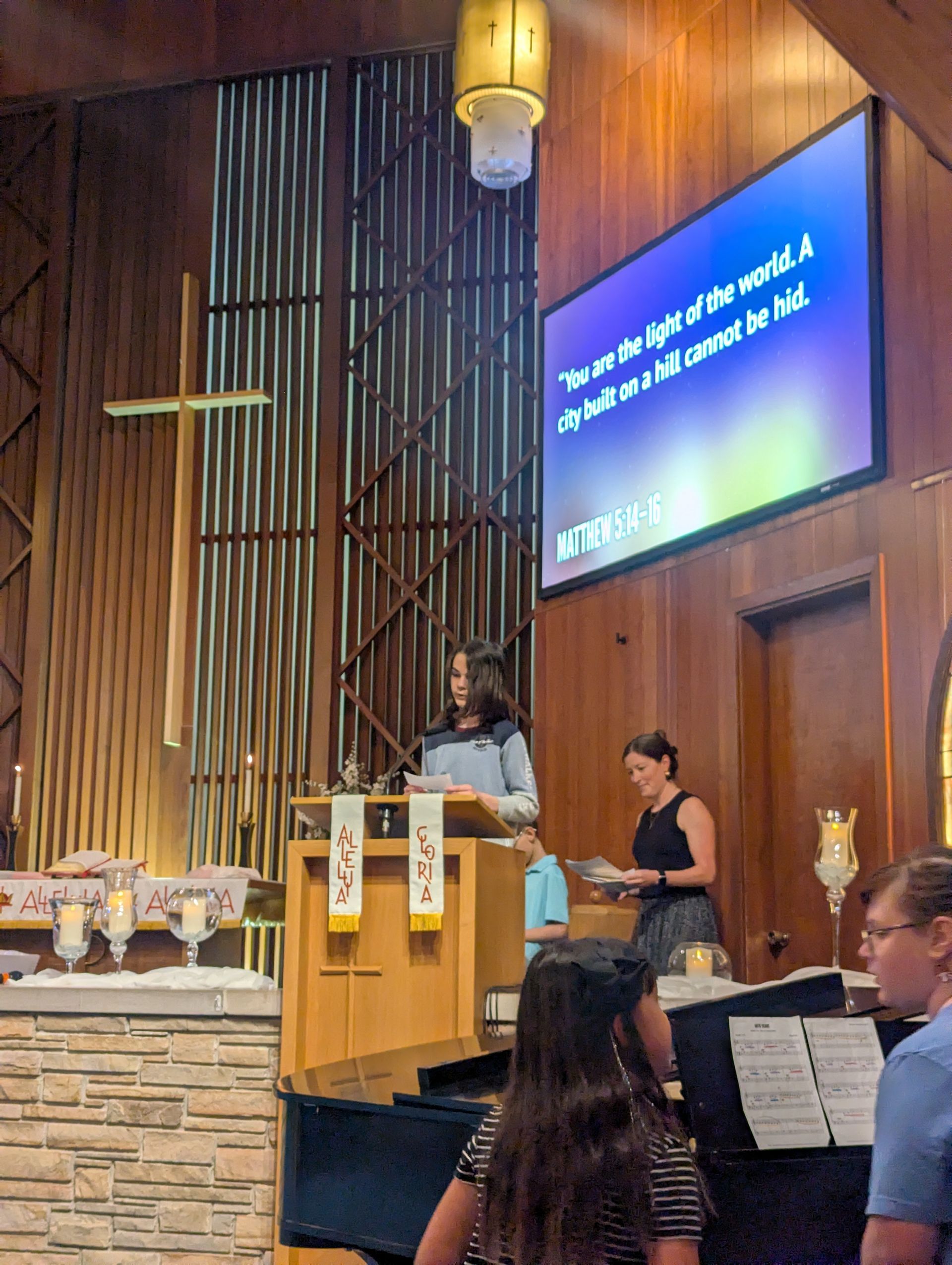 Speaker at podium in wood-paneled church, with projected lyrics and musicians nearby