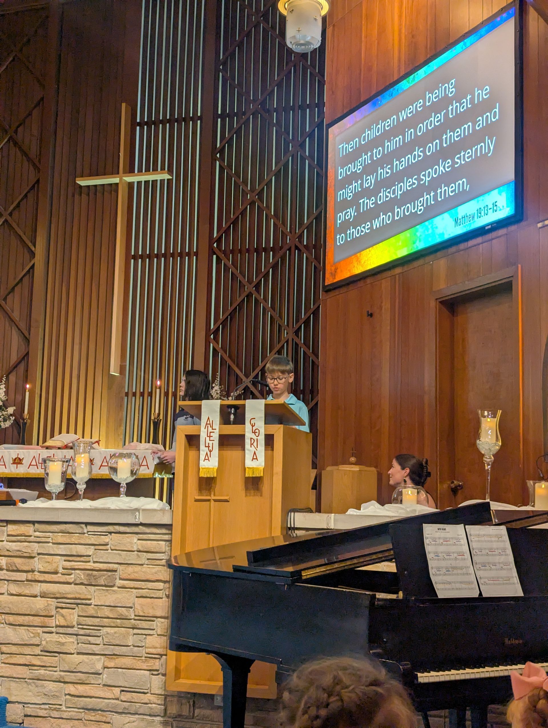 Speaker at church lectern beside a grand piano, with hymn lyrics projected on a screen.