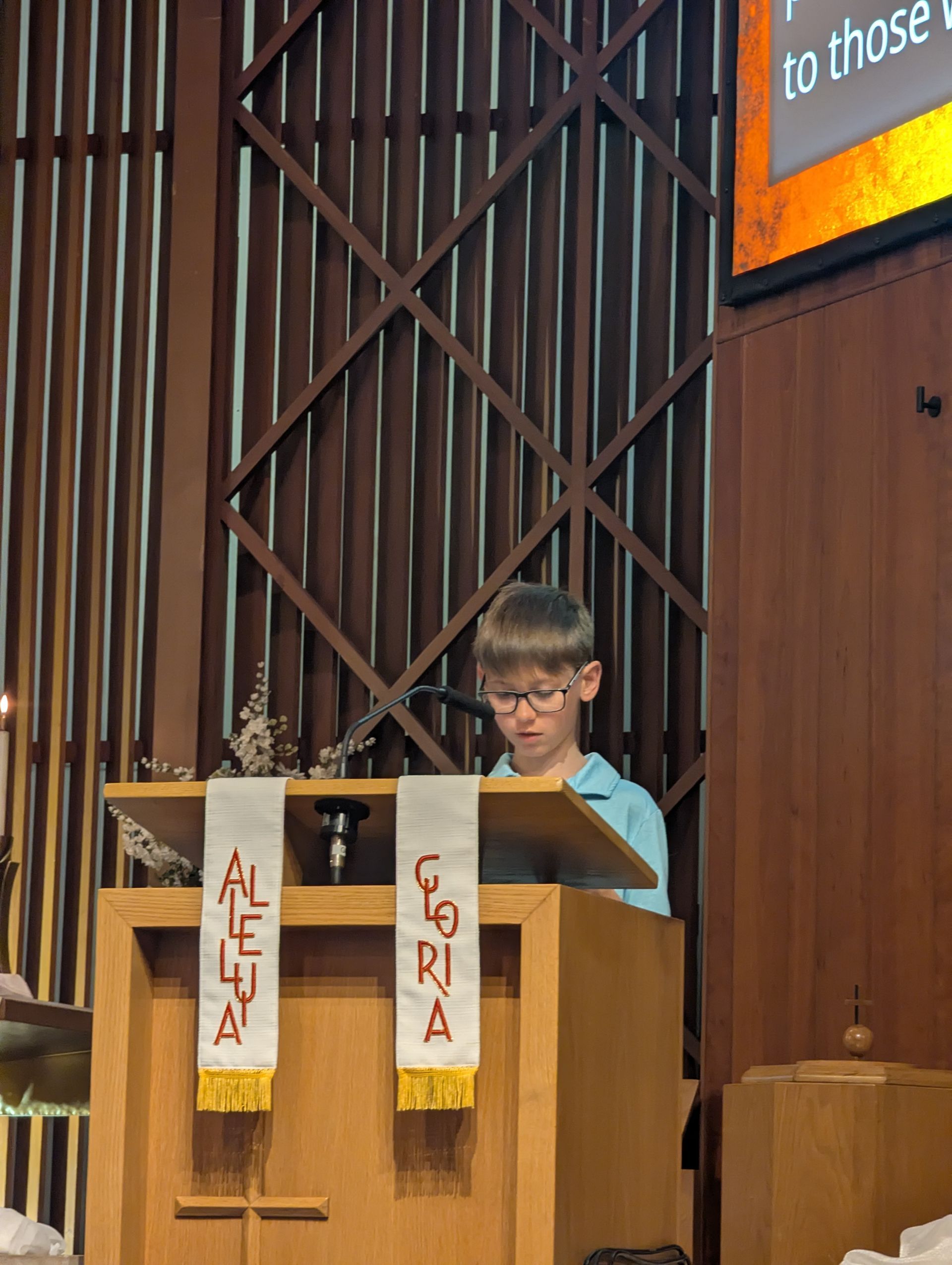 Child reading at a church lectern, with wooden paneling and a slide screen in the background.