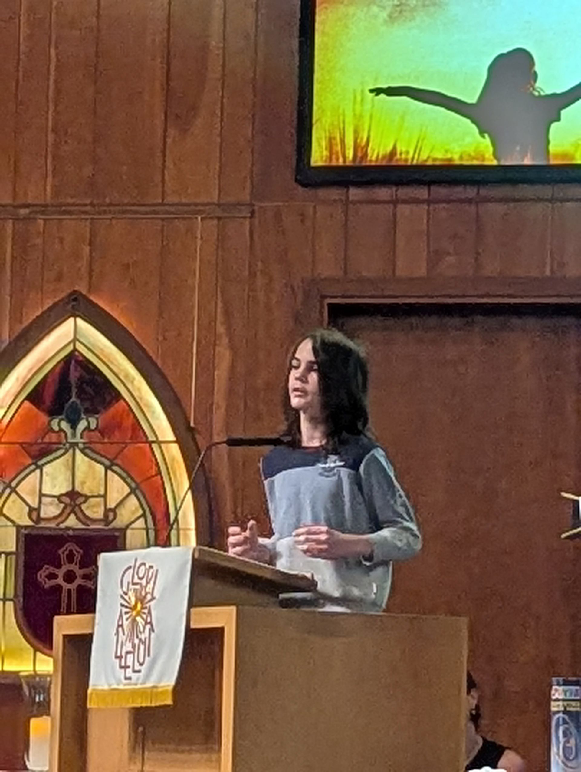 Woman speaking at a church lectern with stained glass and a lit screen behind her