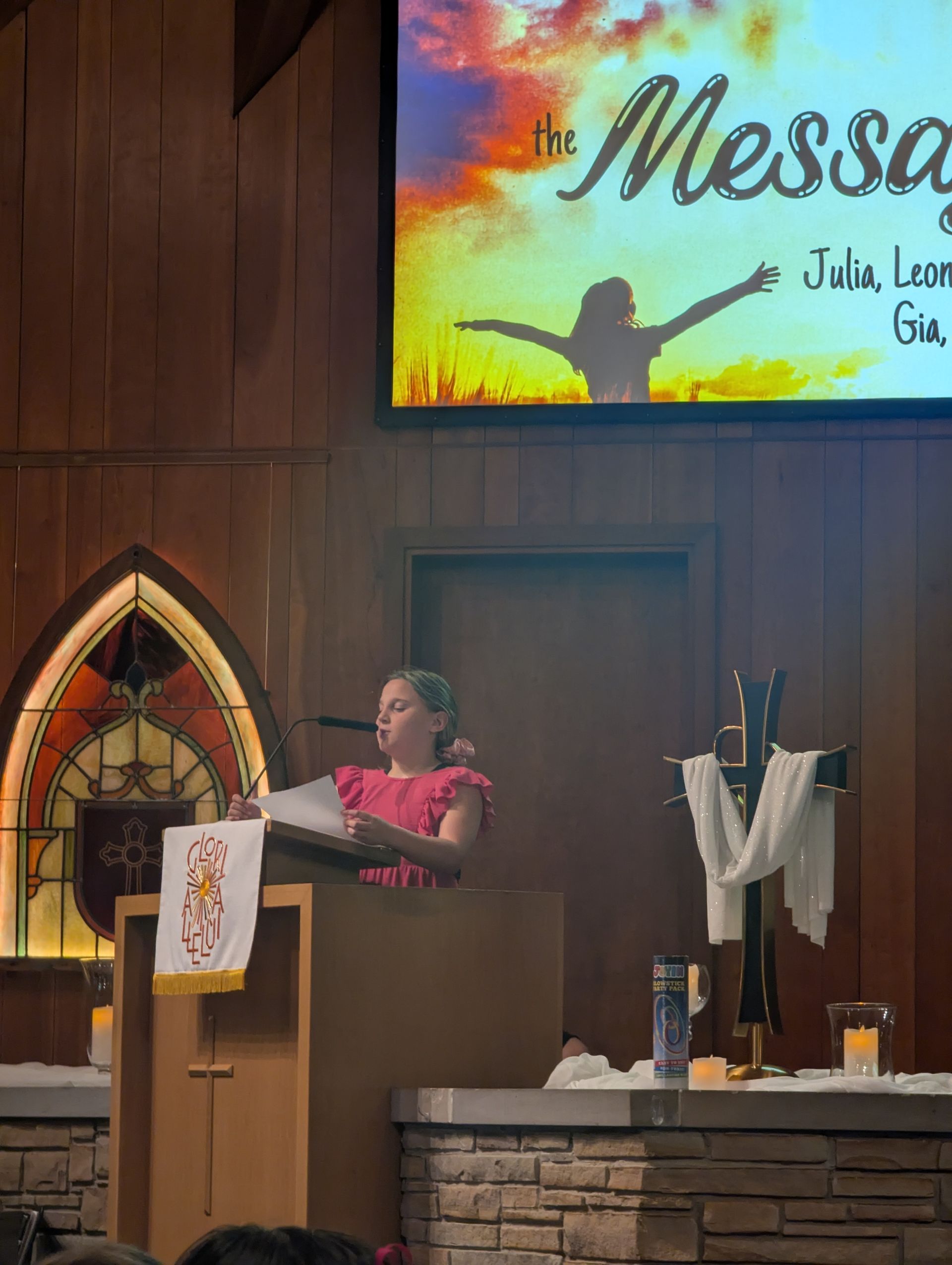 Person speaking at a church pulpit beside a cross, with a stained-glass window and “The Messiah” screen above.