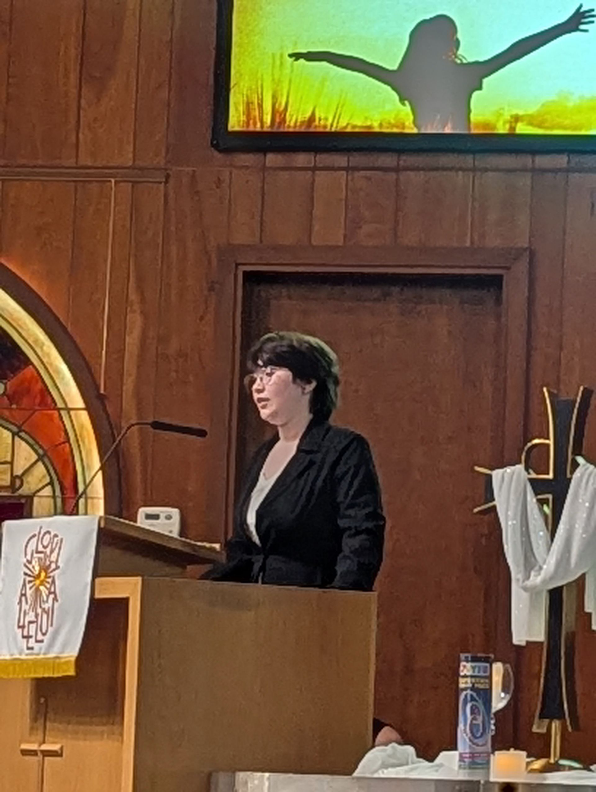 Woman speaking at a church lectern, with a cross banner and a screen showing a silhouetted figure behind her.