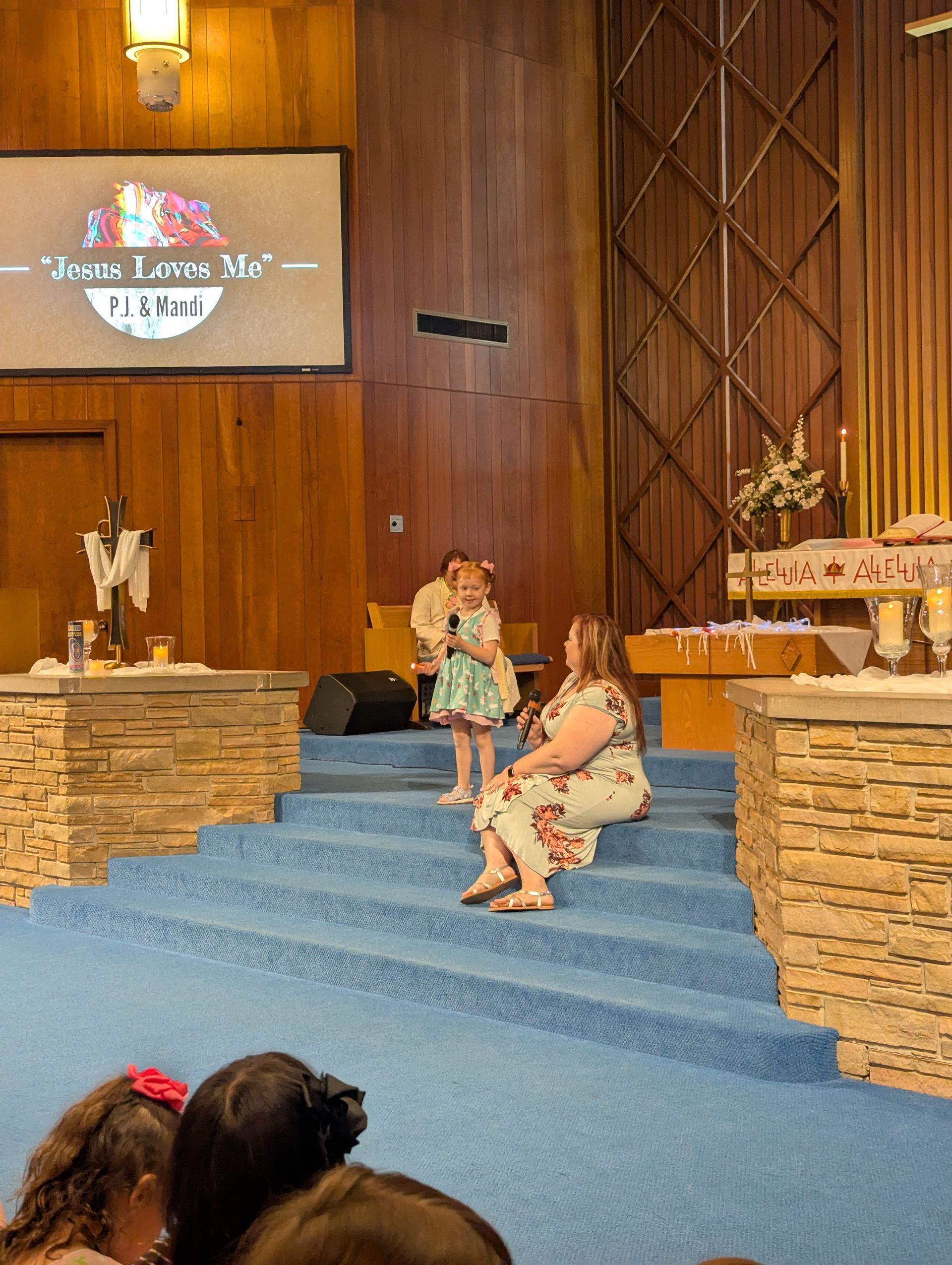Two children sit on church steps facing a stage with a floral backdrop and stone accents.