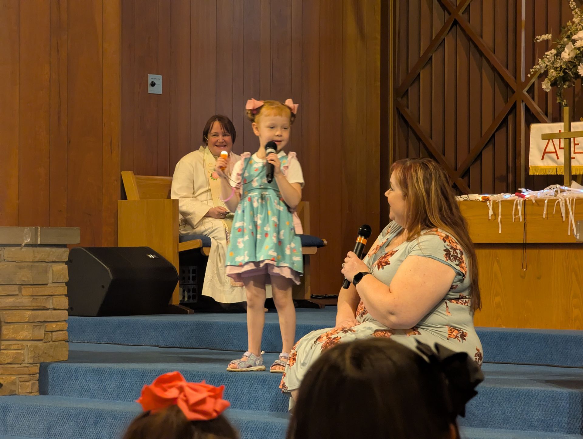 Child in teal dress stands on a stage holding a microphone, with seated audience and wood-paneled backdrop.