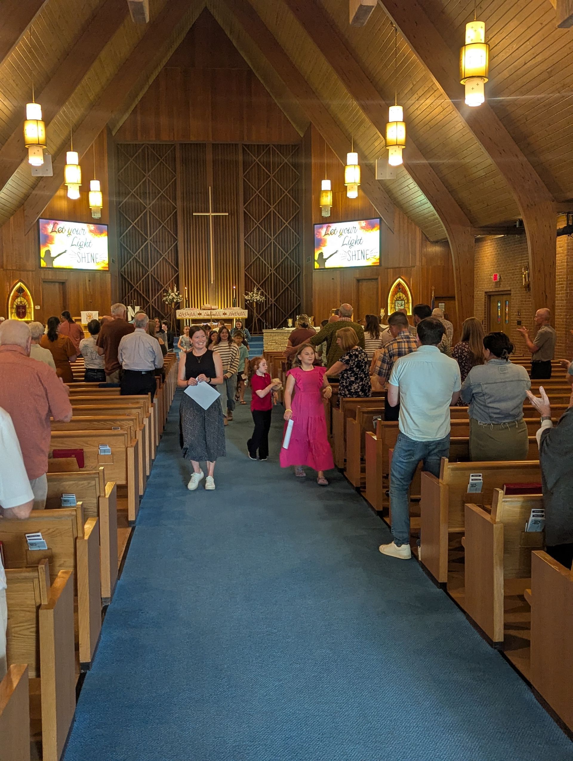 Congregants stand in a wooden church aisle during a service, with stained glass and an altar at the front.