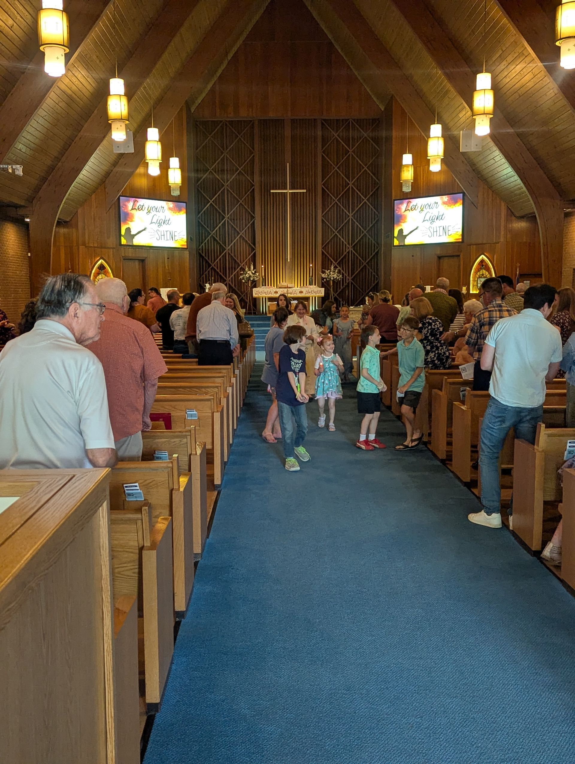 People walking down a church aisle with wooden pews, blue carpet, and warm hanging lights.