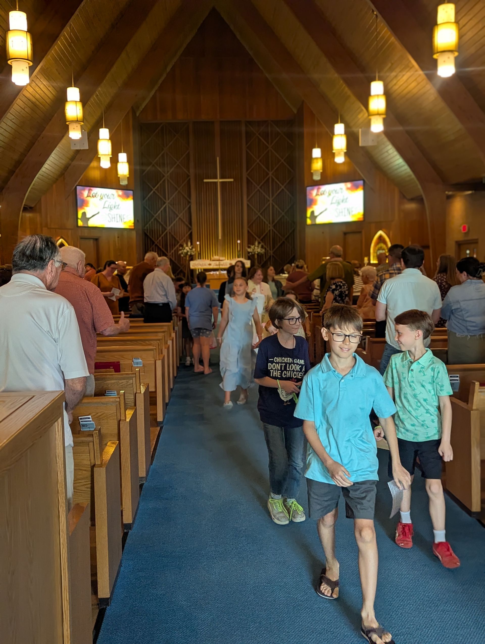 Children walking down the aisle of a church with wooden pews and hanging lights