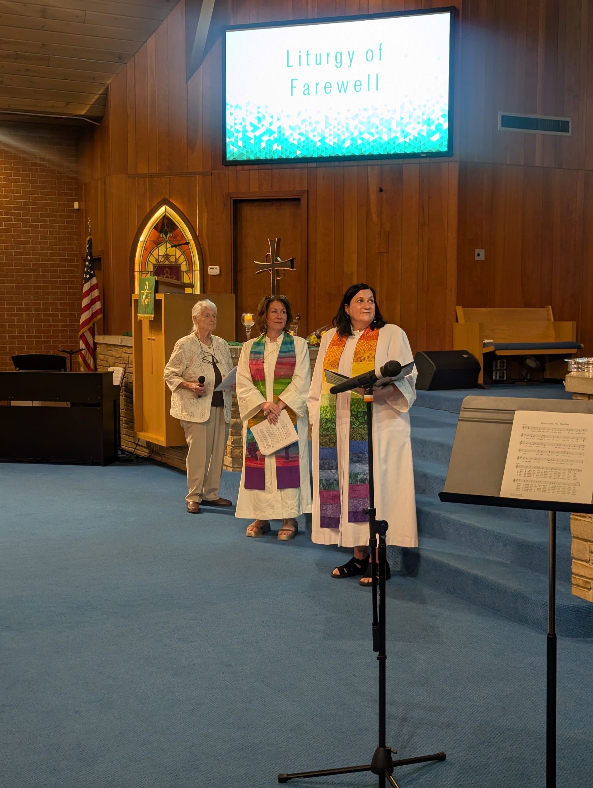 Three people in colorful robes stand in a church sanctuary near a microphone stand.