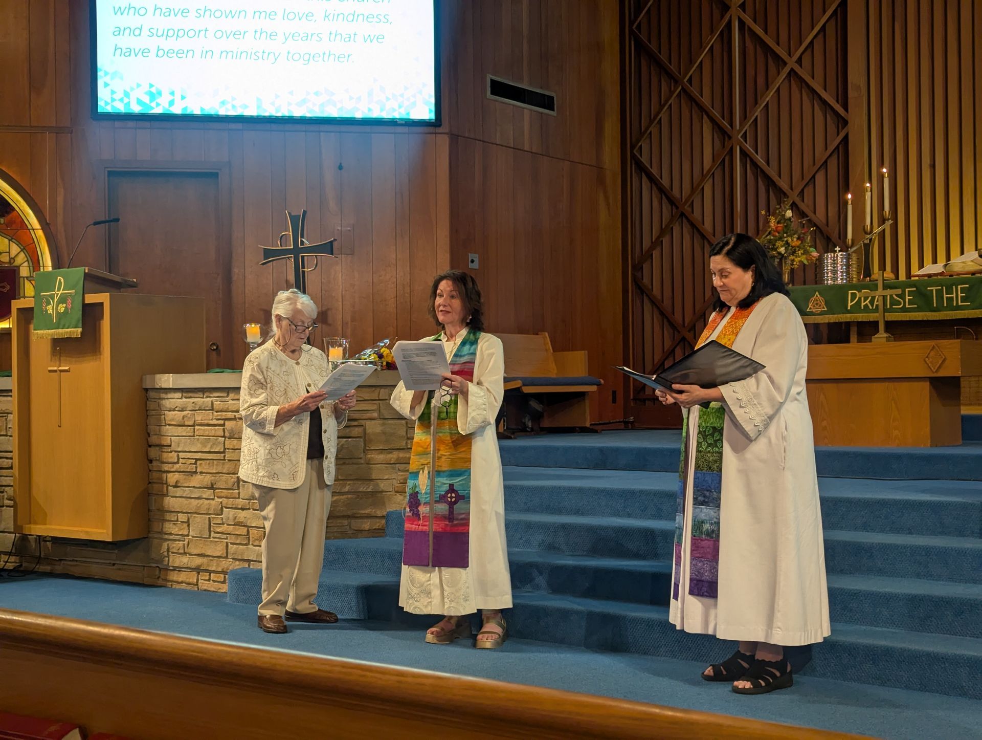 Three people on a church stage, two holding papers, with a cross and podium nearby.