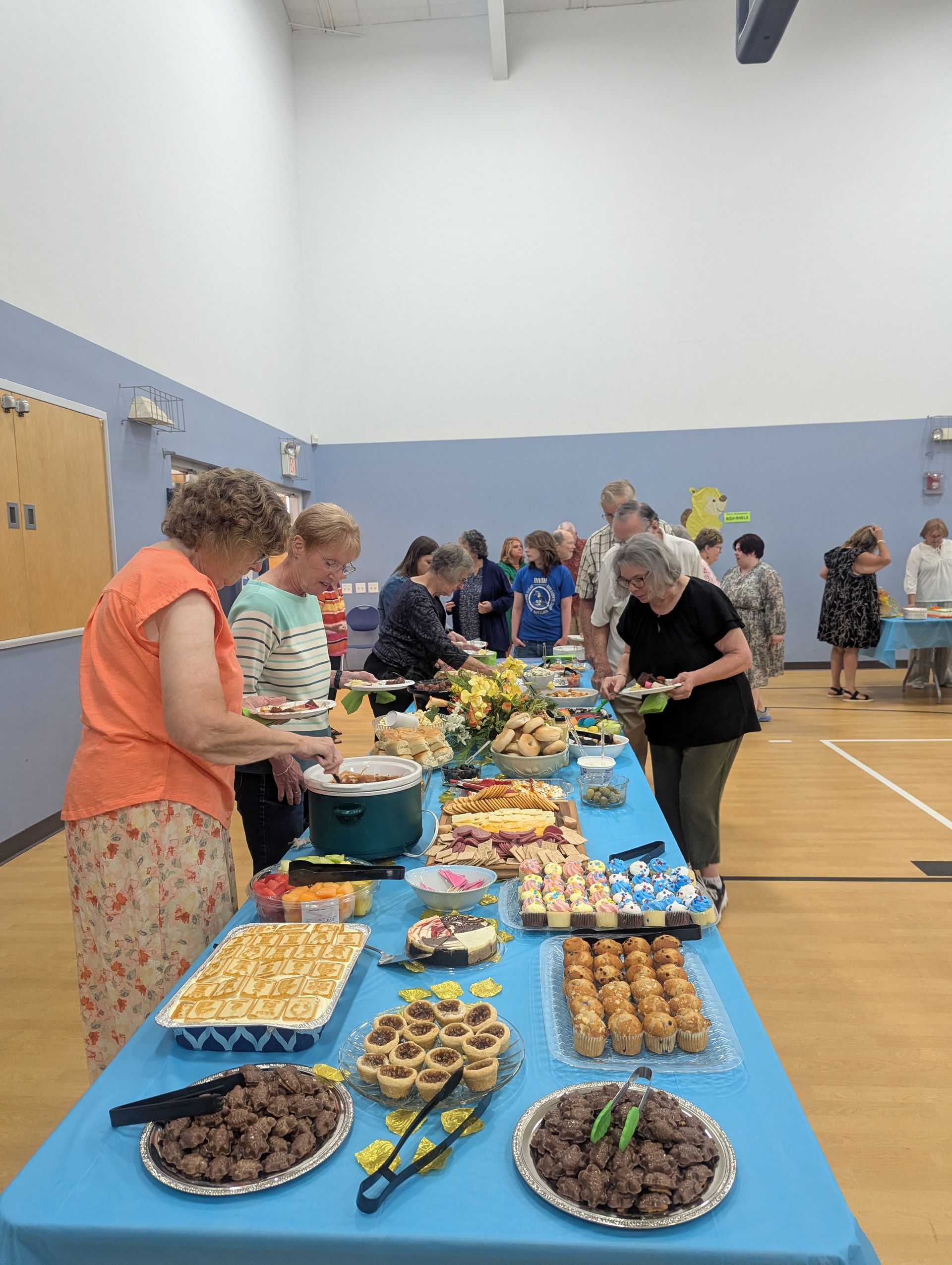 People browsing a long table of assorted baked goods at a community hall event.