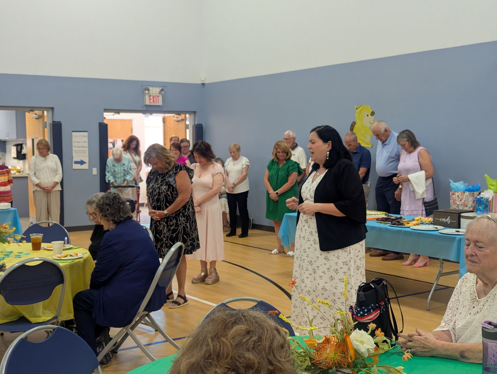 People socializing in a community hall with tables, chairs, and flowers in a blue-walled room.