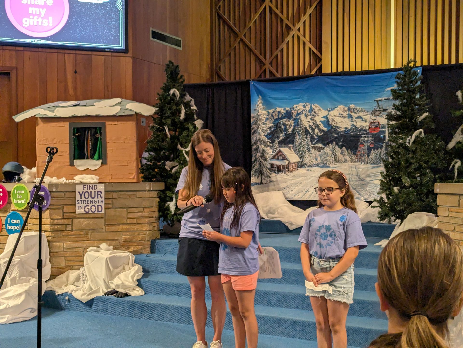 Three children stand on a stage holding papers, with a snowy mountain backdrop and podium nearby.