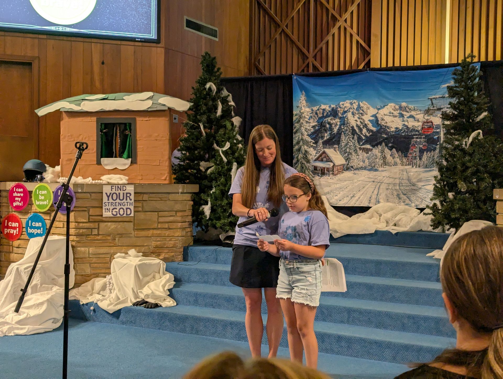 Two children on a church stage holding papers, with a winter mural and small hut backdrop behind them