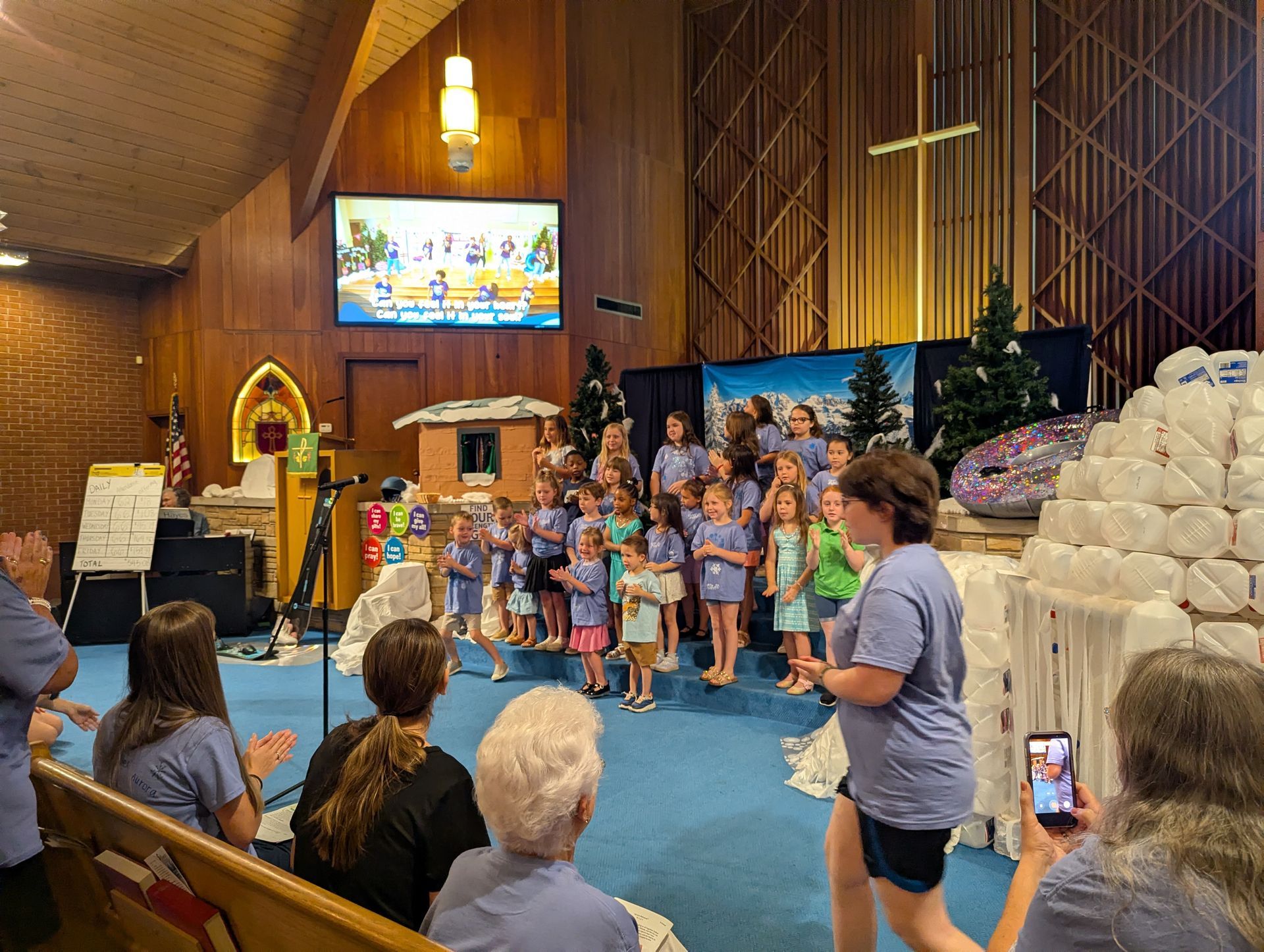 Children performing on a church stage with colorful props and a cross in the background
