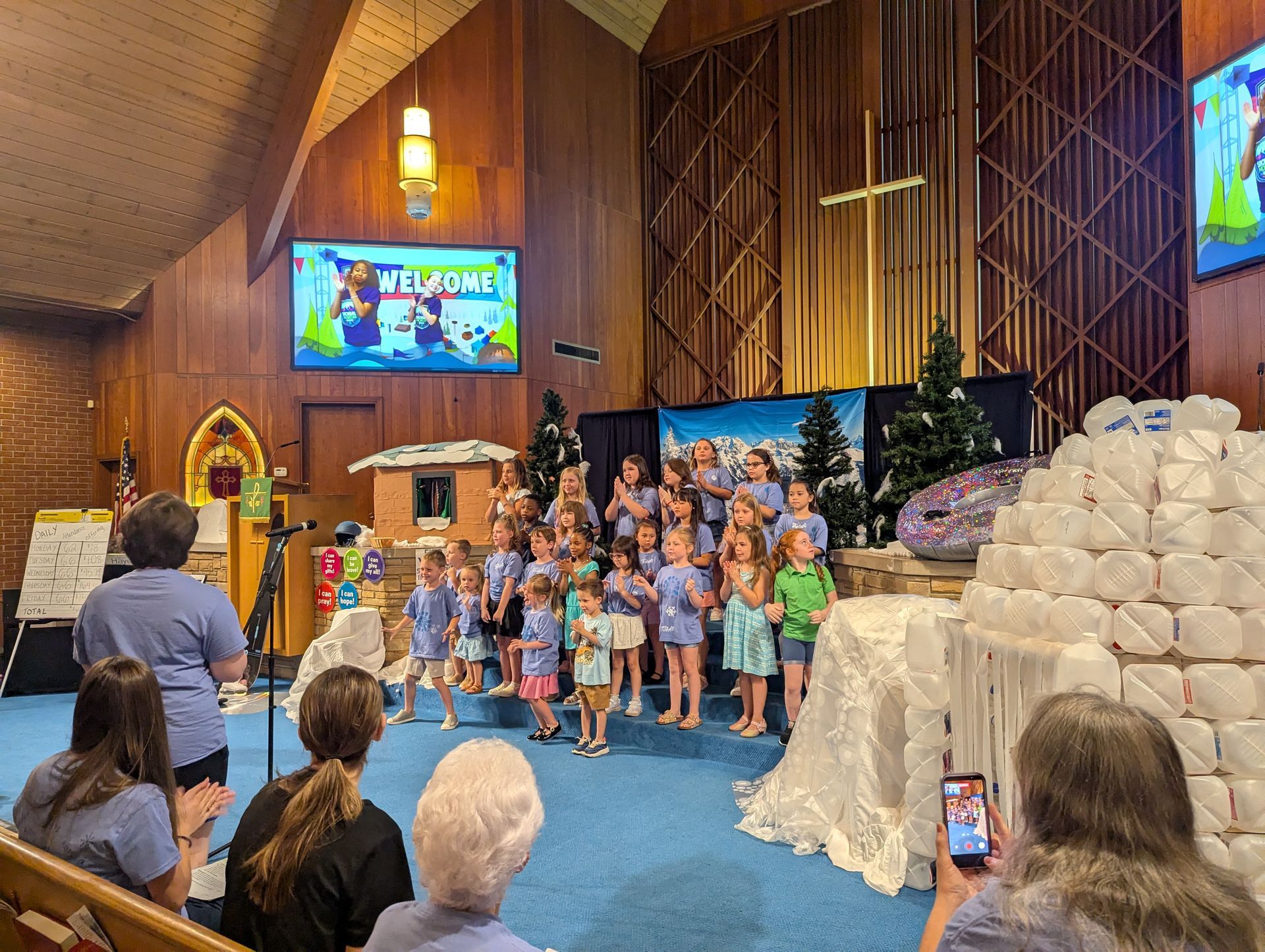 Children posing on a church stage beside a large stack of wrapped gifts with a “Welcome” screen behind them