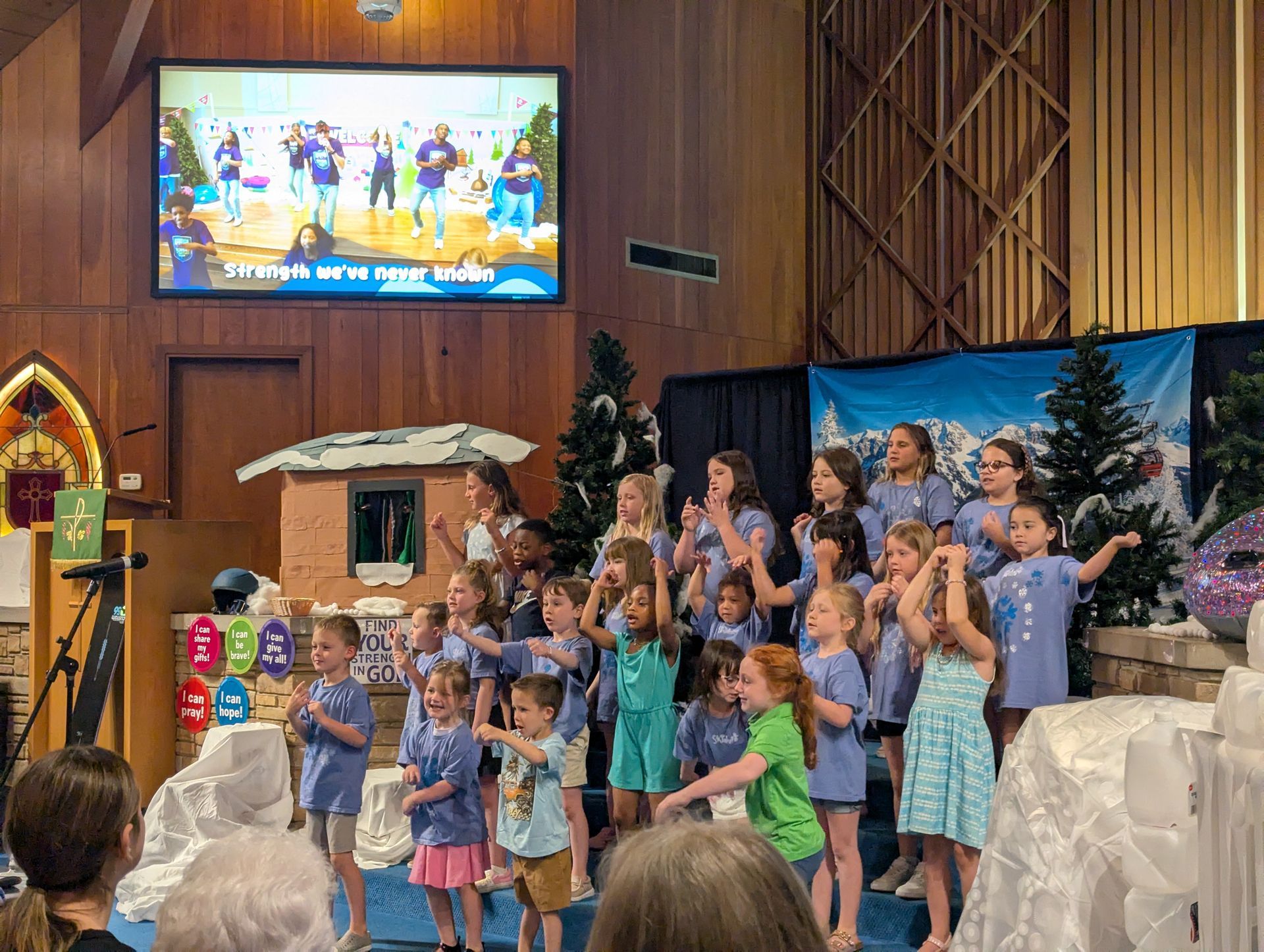 Children in blue and pink costumes performing on a church stage with a nativity backdrop