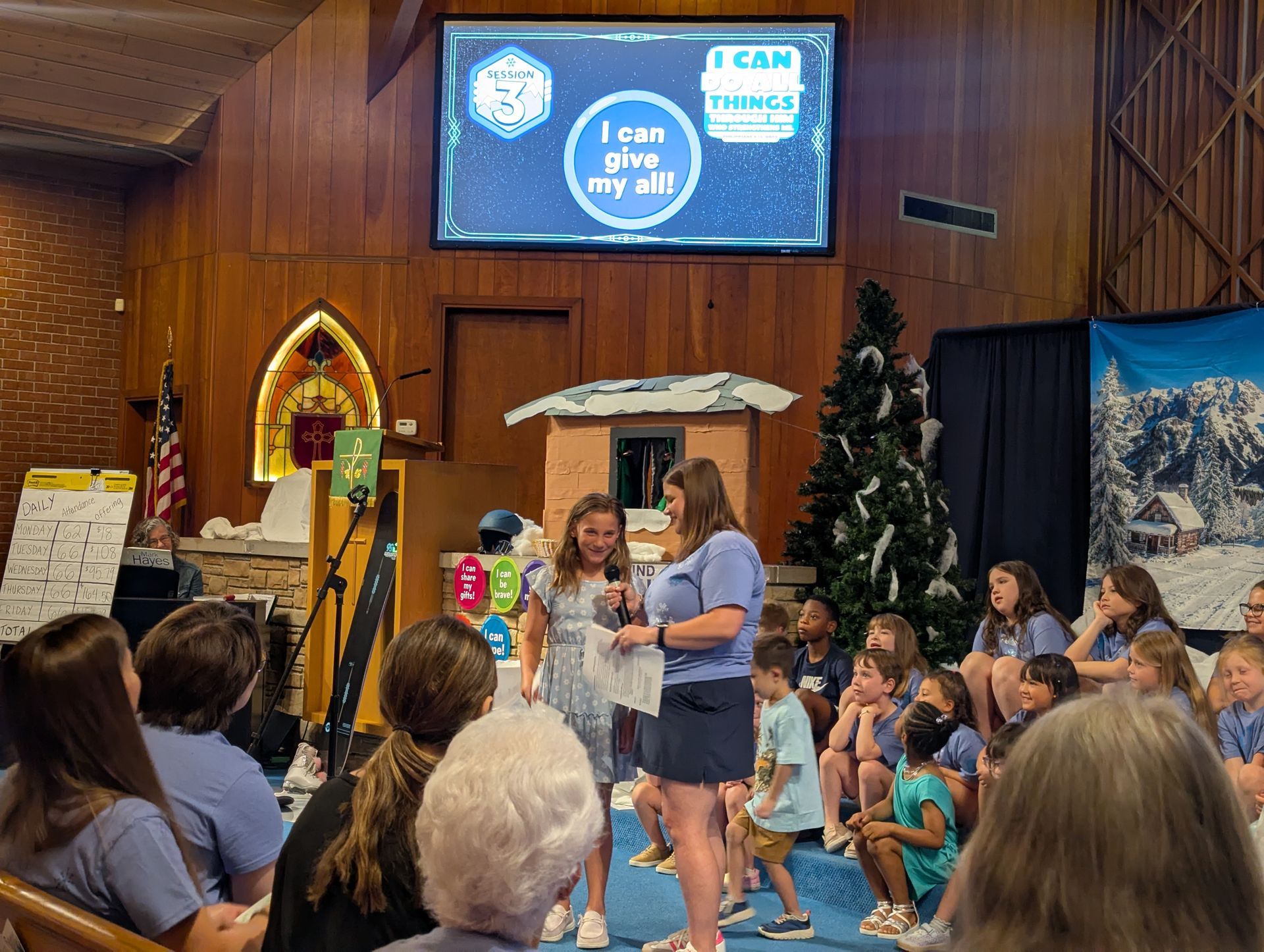 People gathered in a church hall for a children’s event, with a speaker on stage and a screen behind her.