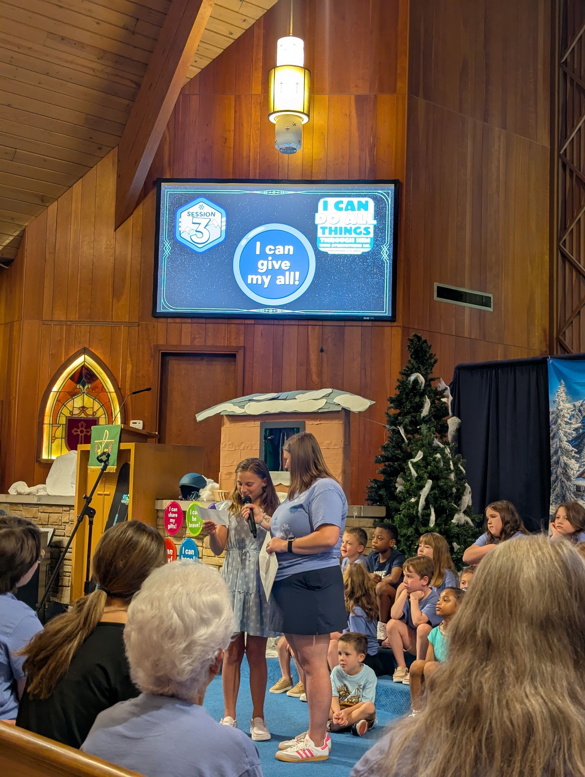 Woman speaking to children in a church hall during a holiday event, with Christmas tree and nativity display.