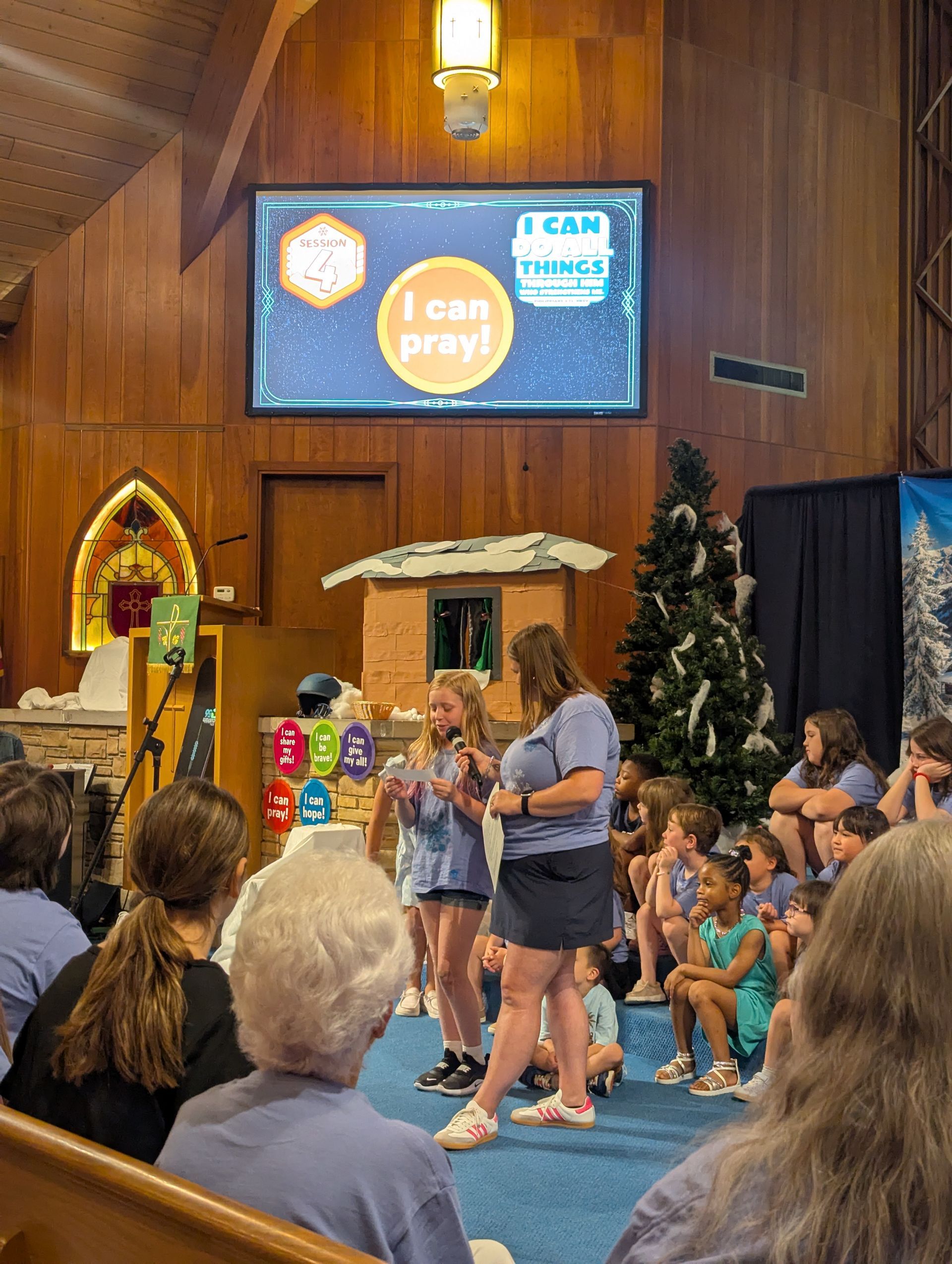 Children and adults gathered in a church hall for a prayer event with a screen displaying “I can pray.”