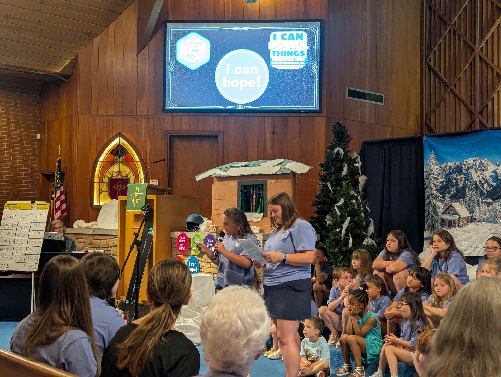 People seated in a church hall watch a woman speaking at a podium beside a screen and Christmas decorations.