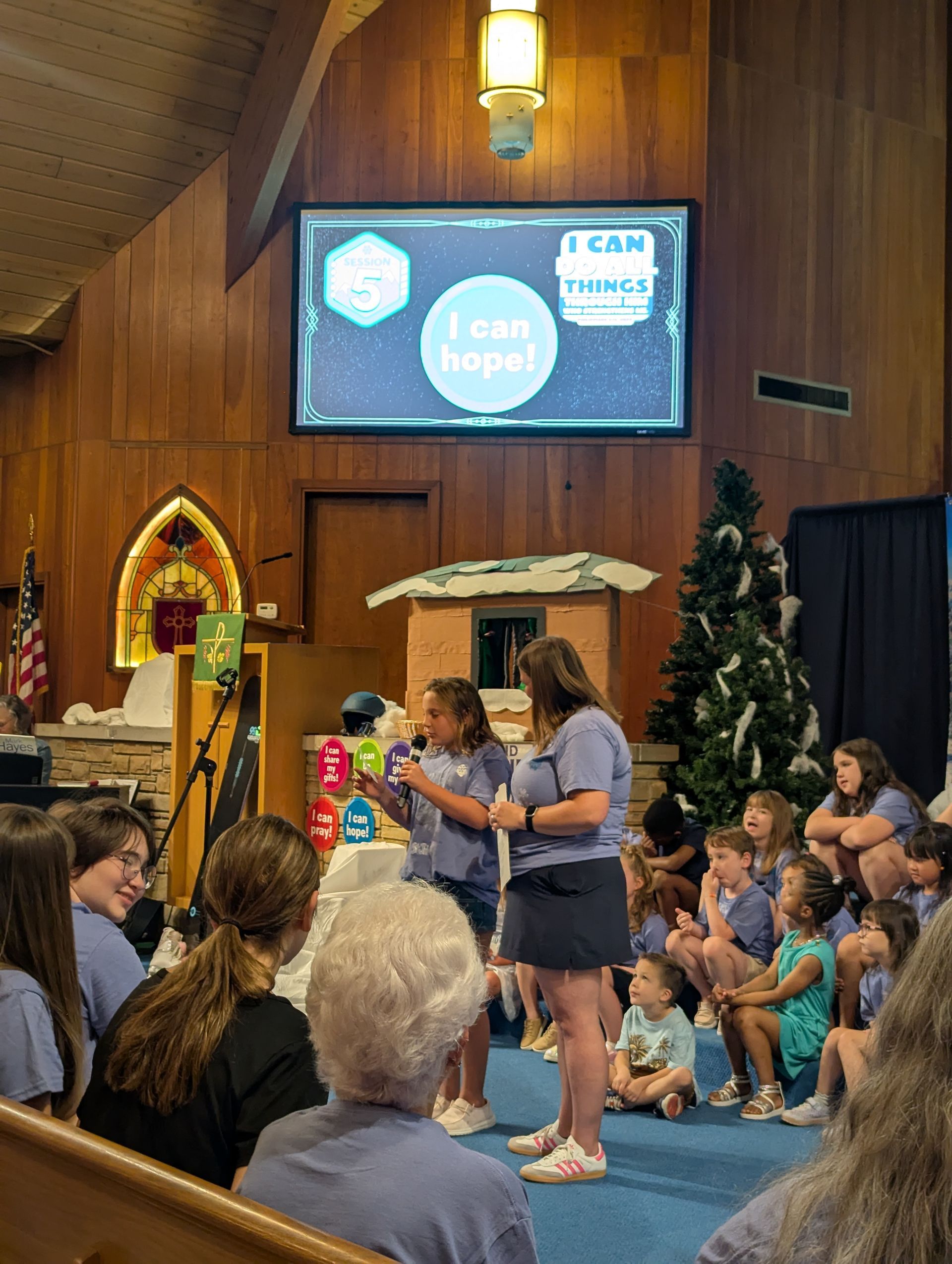 Children seated in a church, watching a woman speak on a stage with a screen behind her.