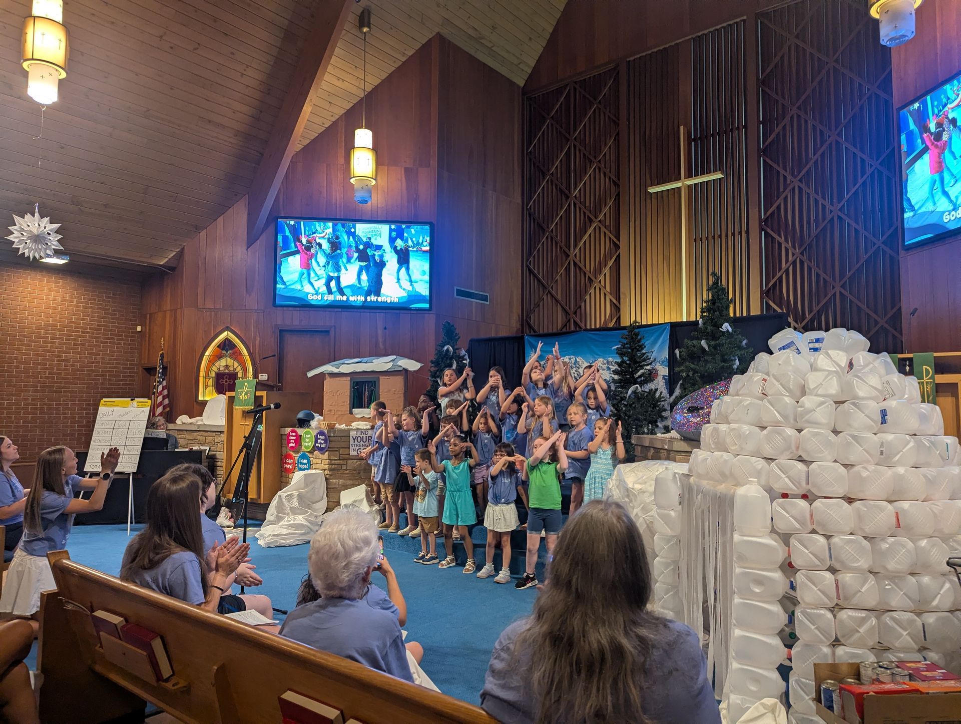 Children performing in a church sanctuary beside a pyramid of white cups, with seated audience members watching.