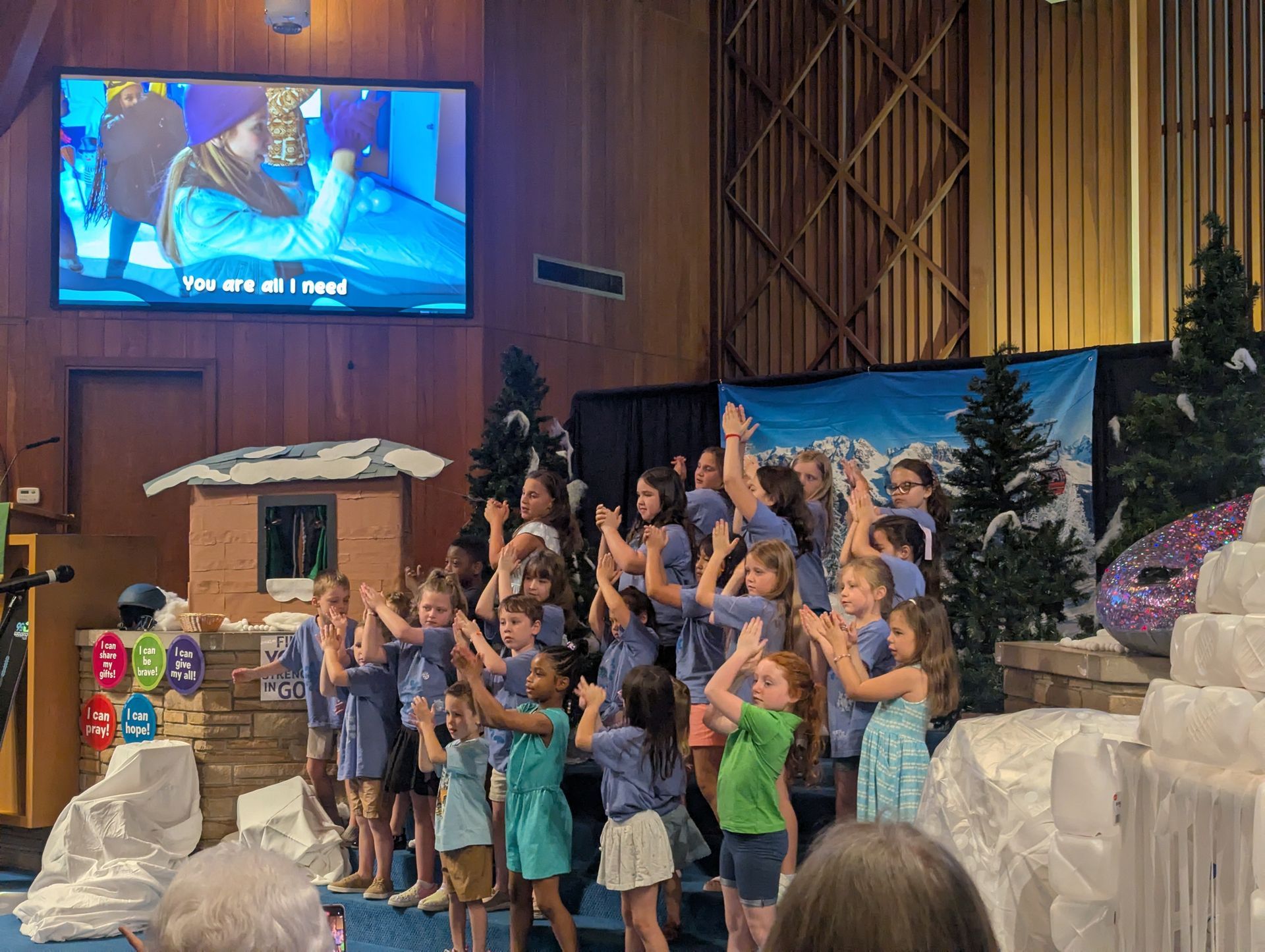 Children performing on a church stage with a large screen, blue backdrop, and white set pieces