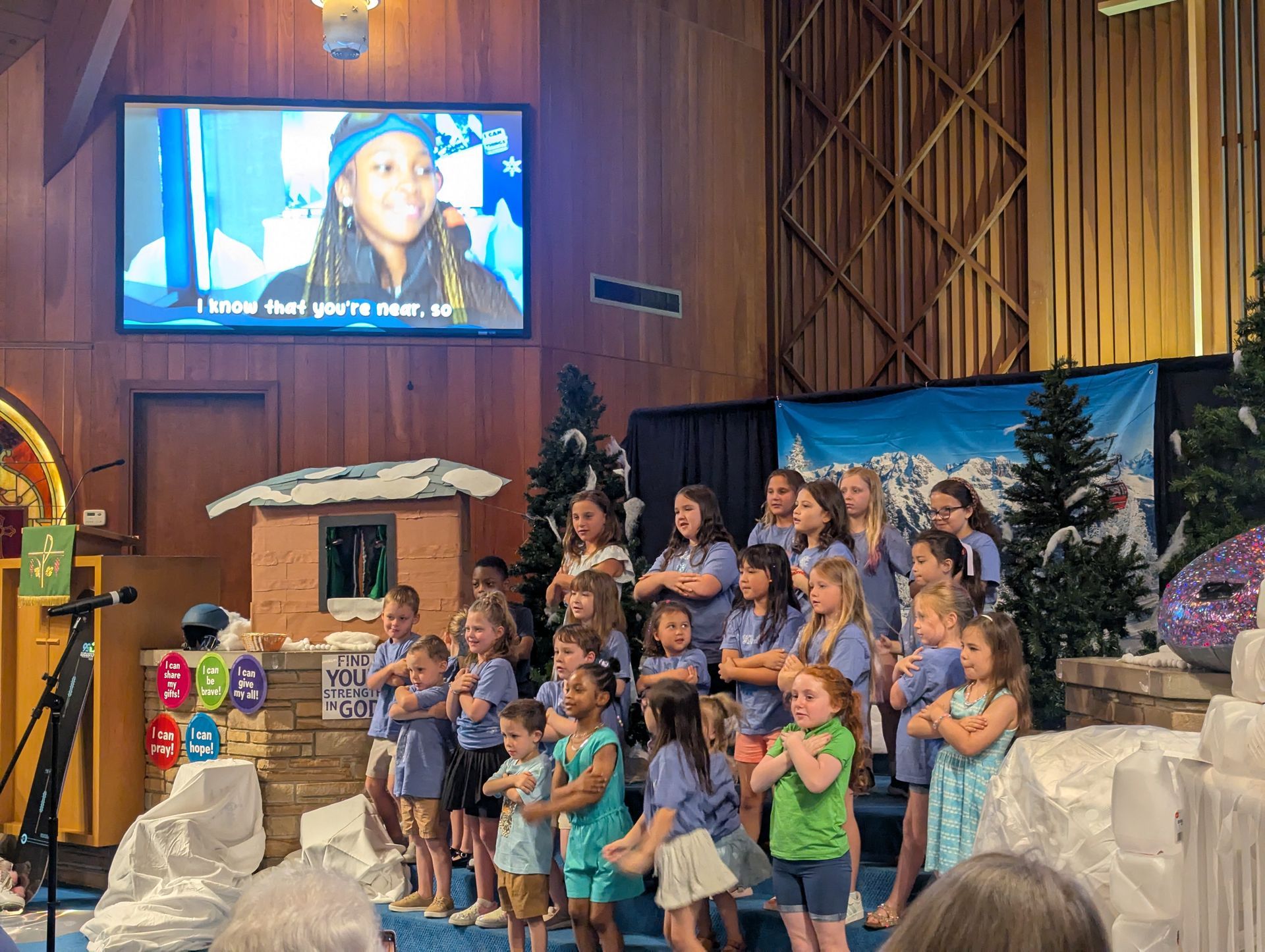 Children singing on a church stage beside a screen showing a smiling child in a winter hat