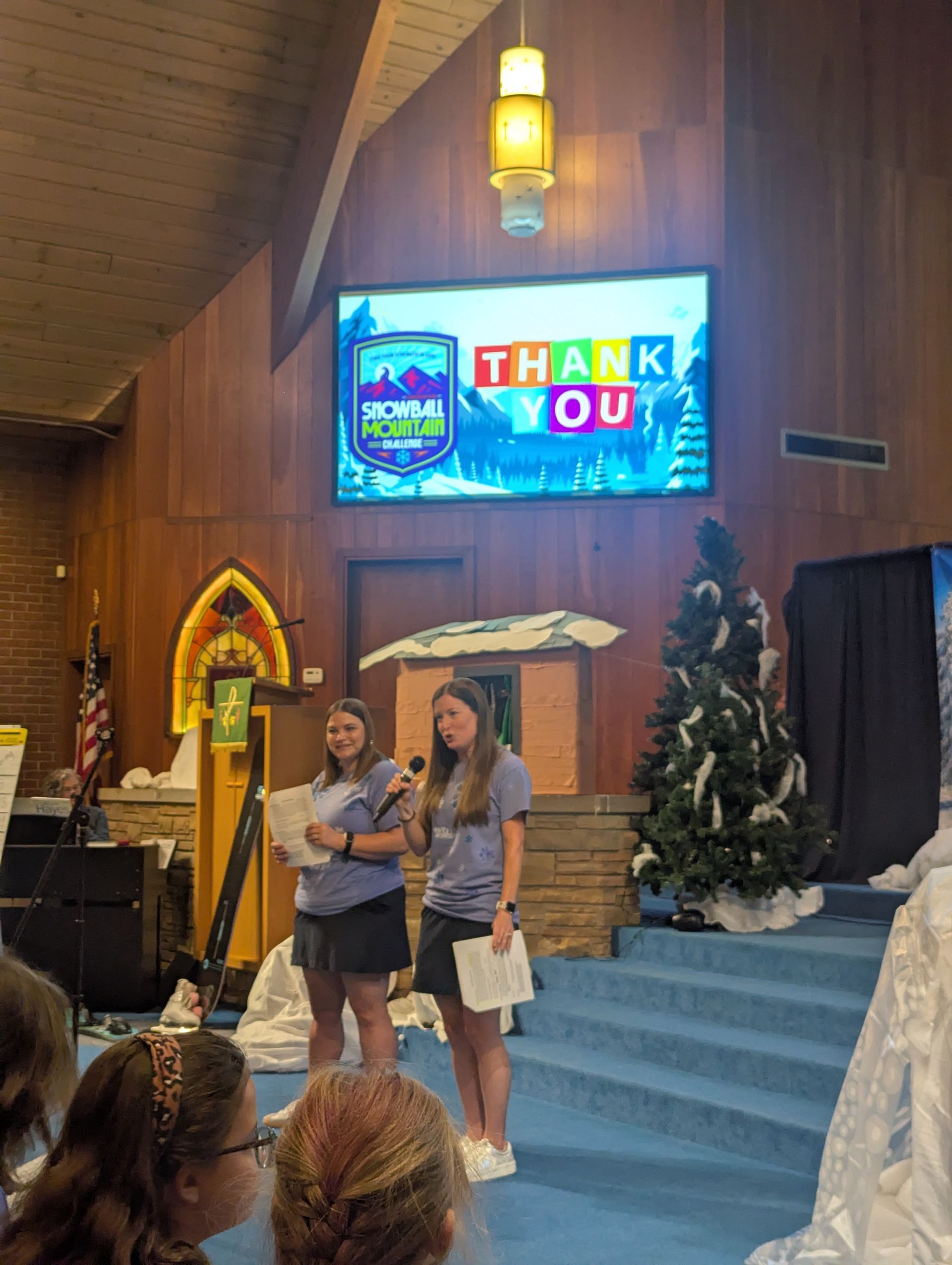Two people stand on a church stage under a “Thank You” screen, with steps, candles, and a Christmas tree.