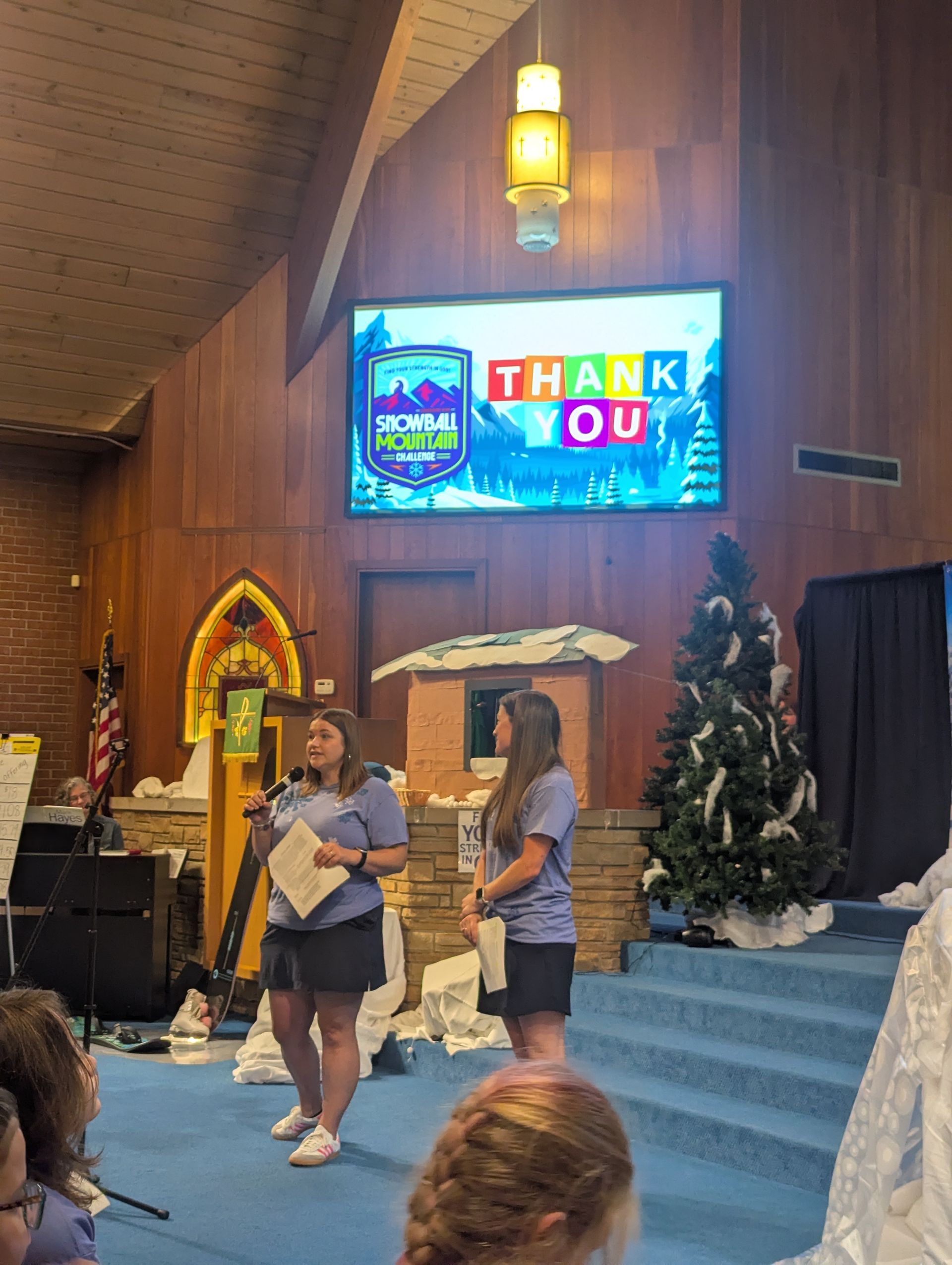 Two women speaking on a church stage beneath a “THANK YOU” screen and Christmas tree decorations