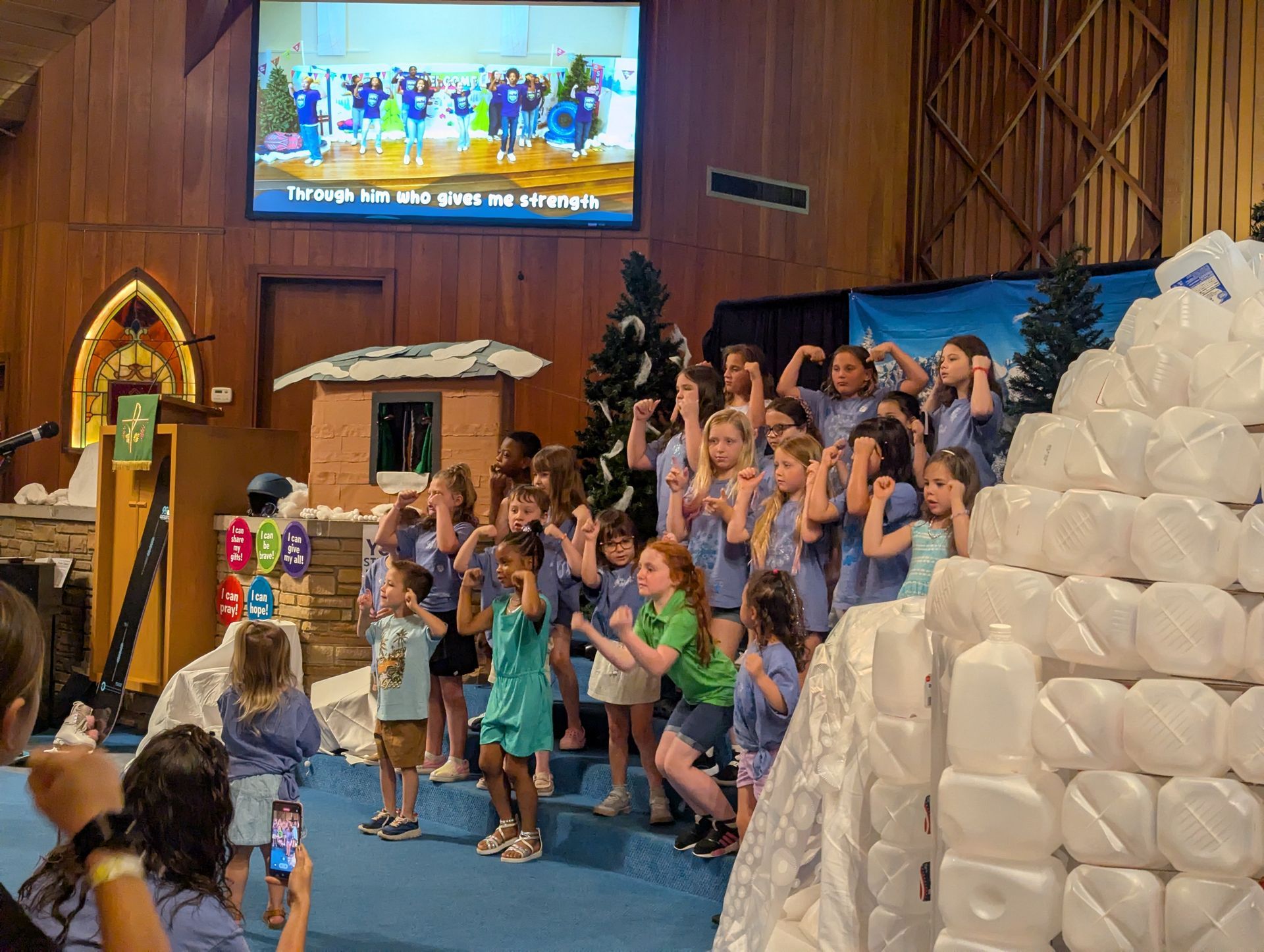 Children sing on a church stage beside a large white stacked display, with a screen and festive decor behind them.