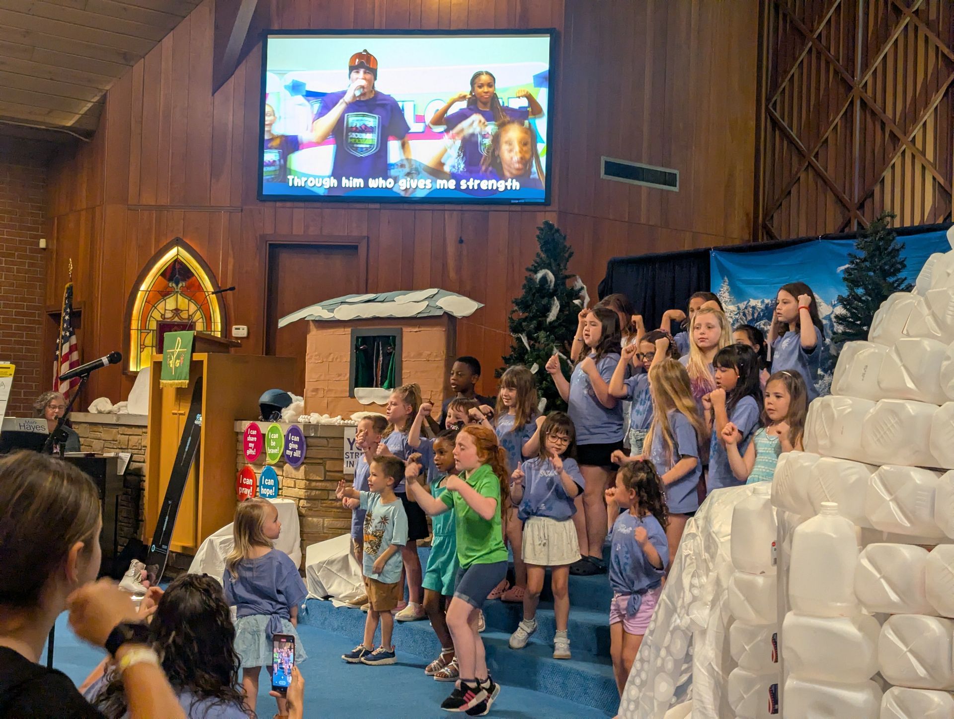 Children on a church stage with raised hands during a group activity, with a large screen behind them.
