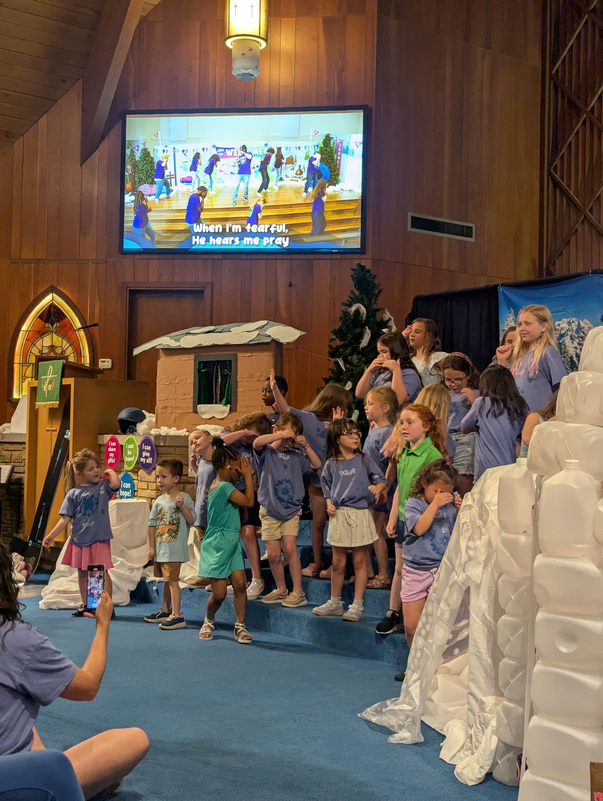 Children standing on church steps during a performance, with a screen overhead and a nativity scene nearby