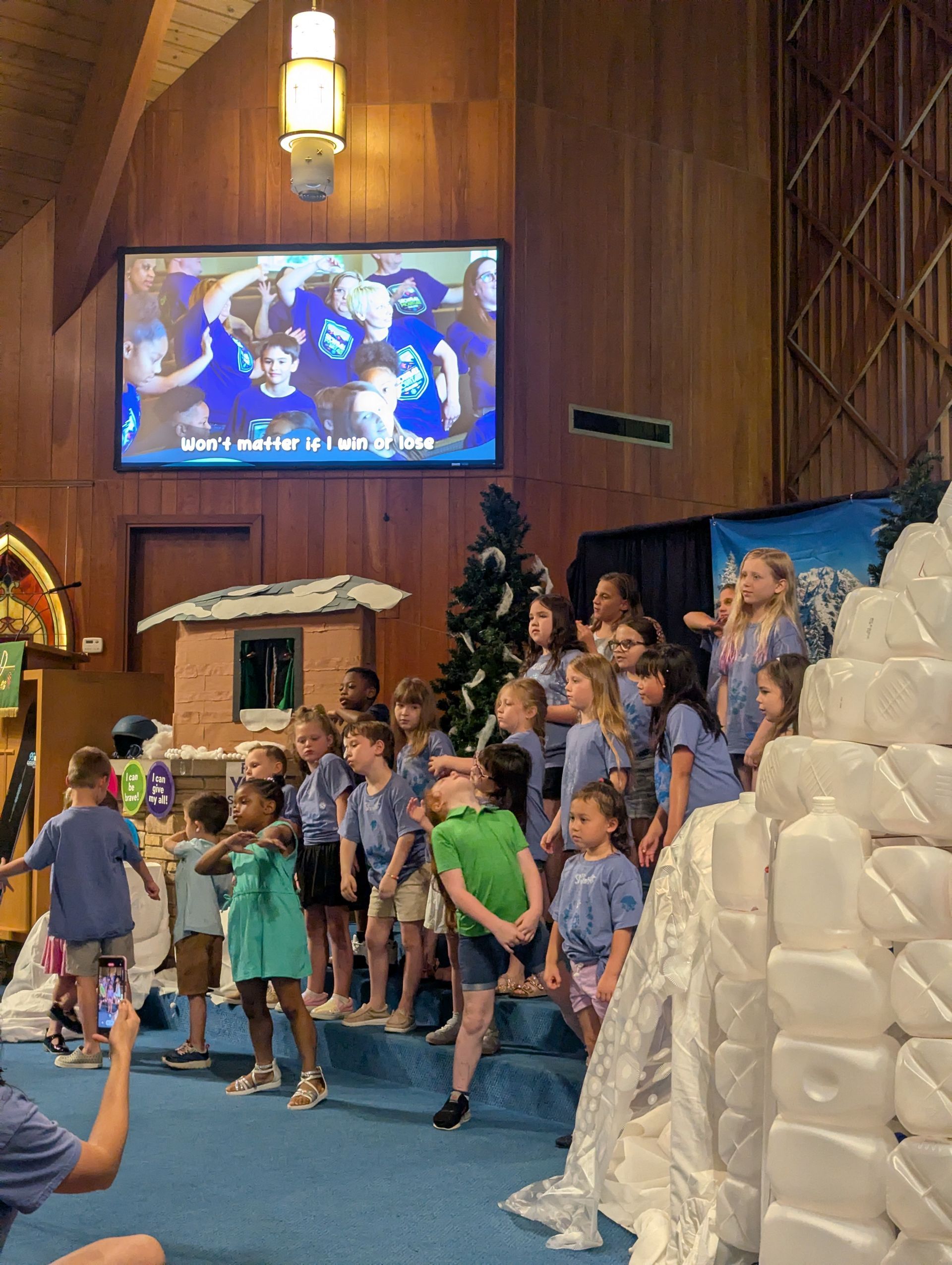 Children singing on a church stage with a large screen behind them and a paper-mâché nativity scene beside them.