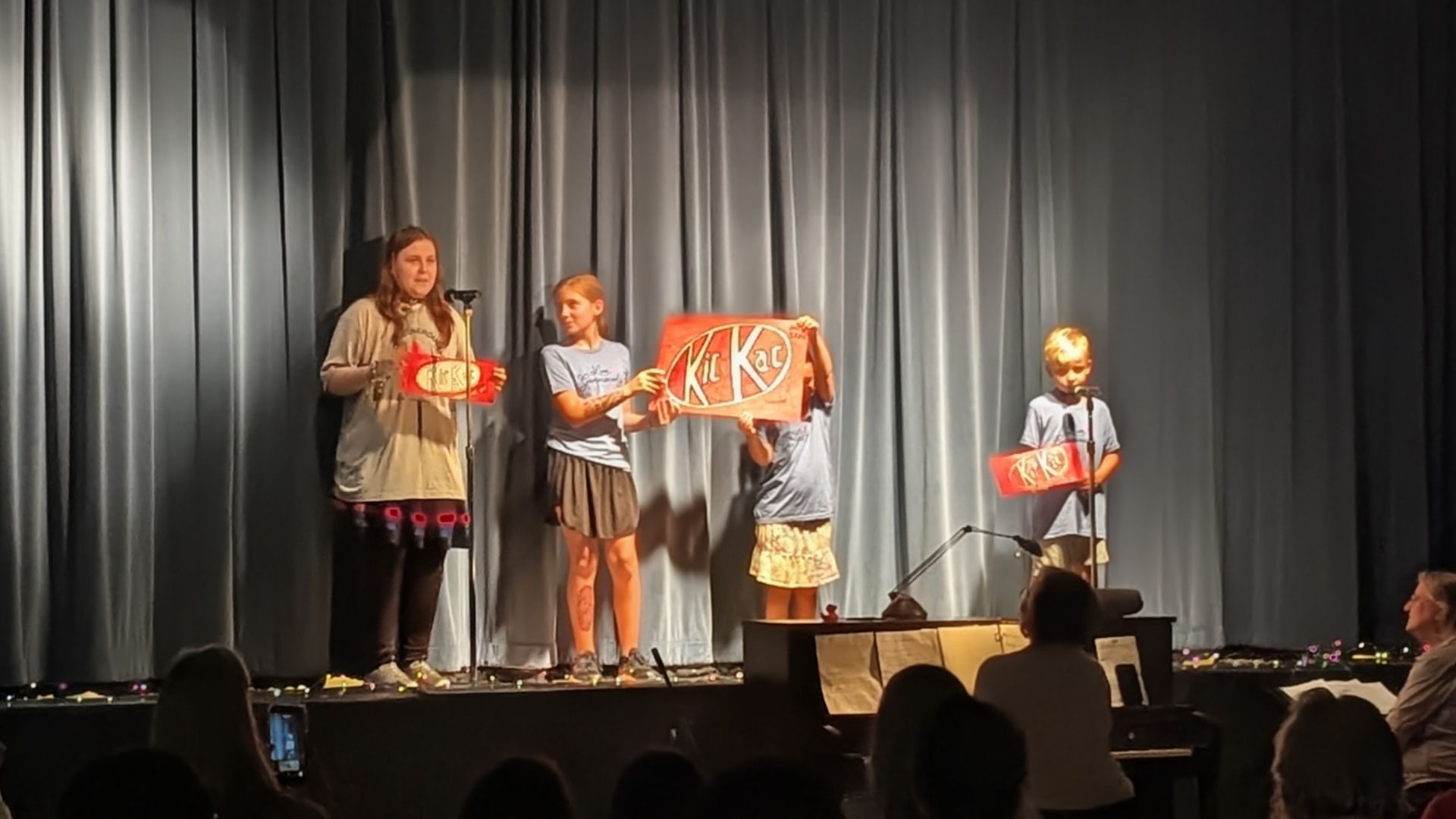 Four children on a stage holding decorated gift boxes in front of silver curtains, with audience silhouettes below