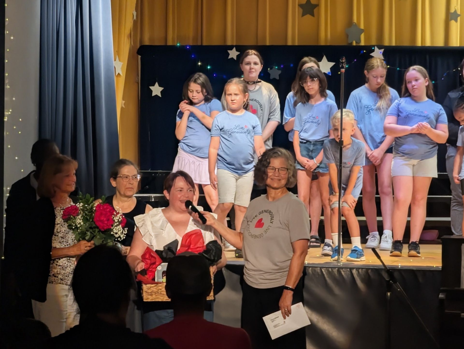 People on a stage holding flowers, with a group of children standing behind them and star decorations in the background.