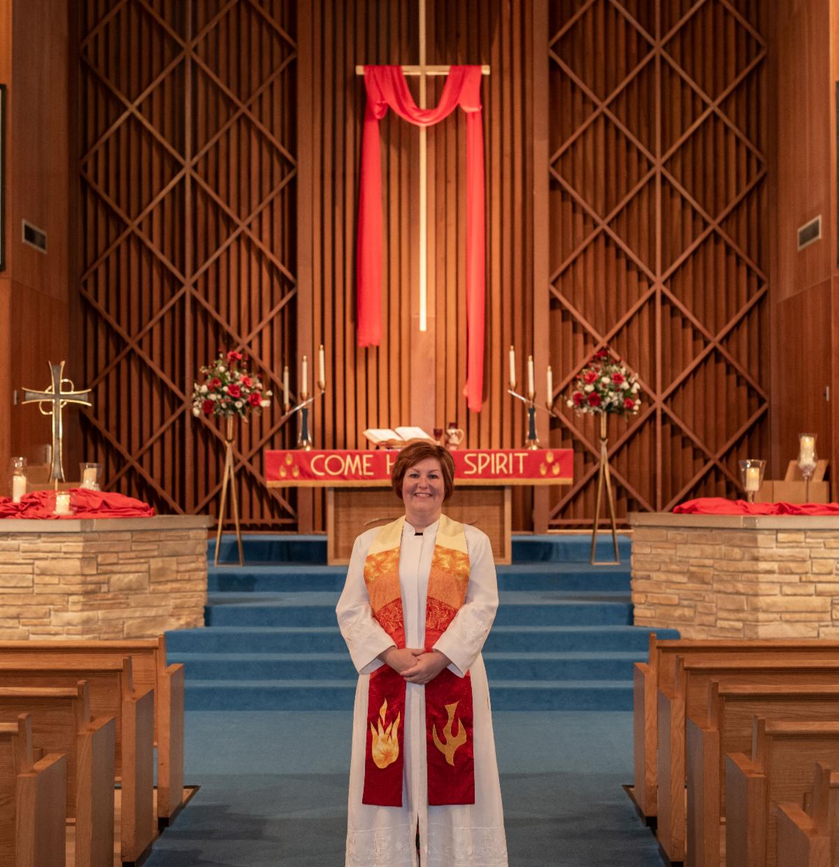 Person in white robe and red stole stands in a church sanctuary before a cross and floral arrangements.