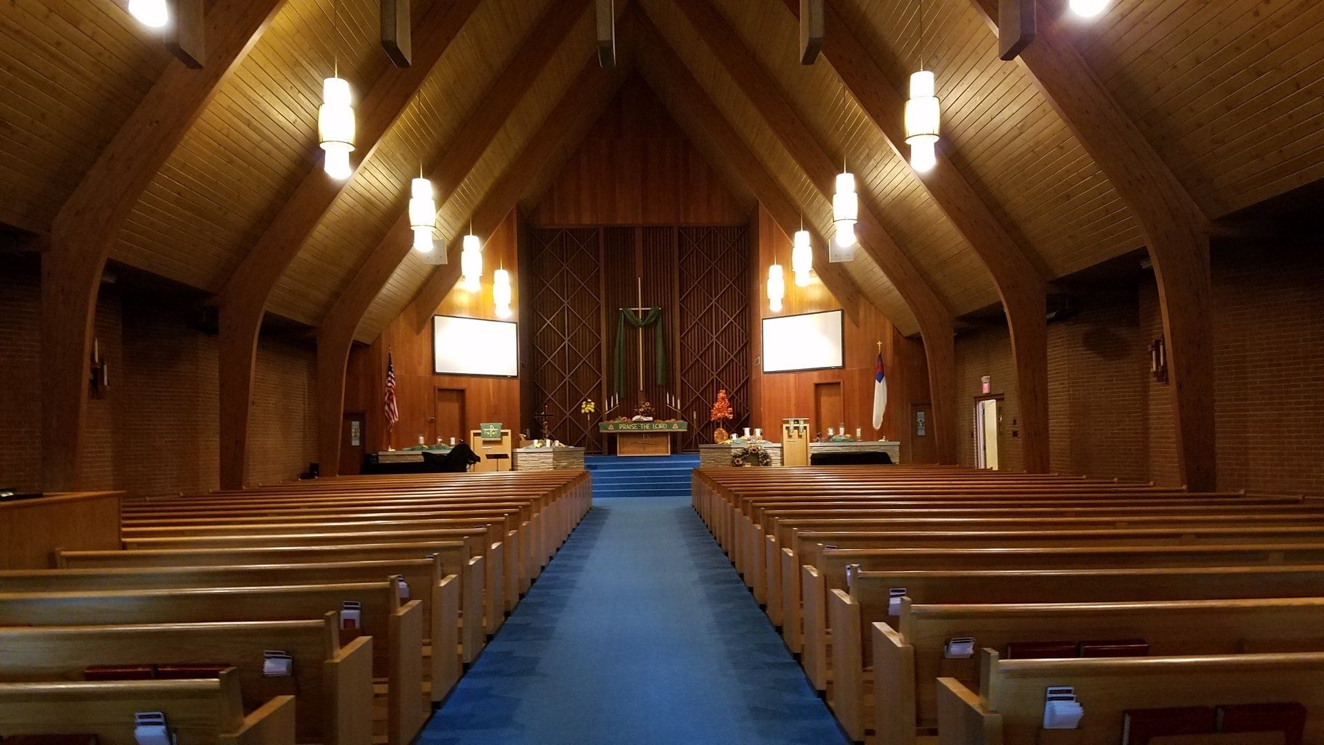 Empty church interior with wooden pews, blue aisle carpet, and hanging lights facing the altar