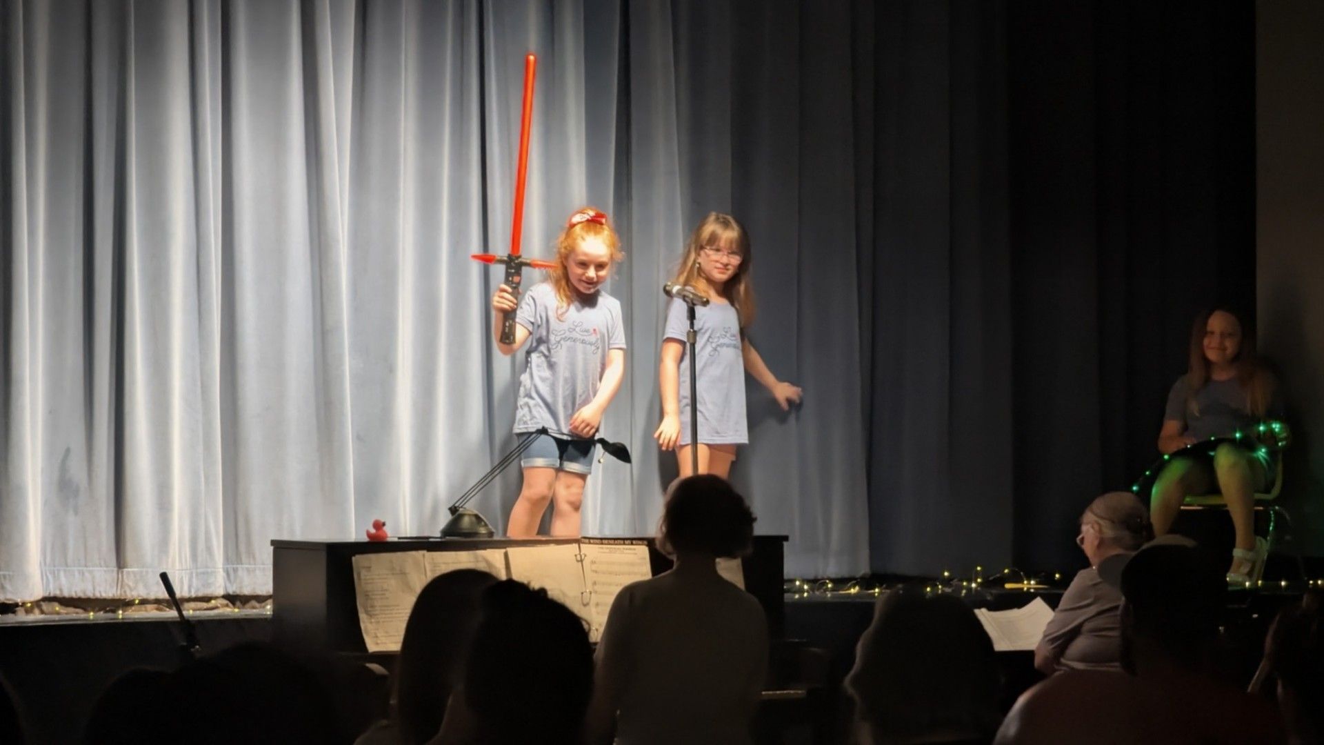 Two children perform onstage with a red staff, in front of a curtain and seated audience.
