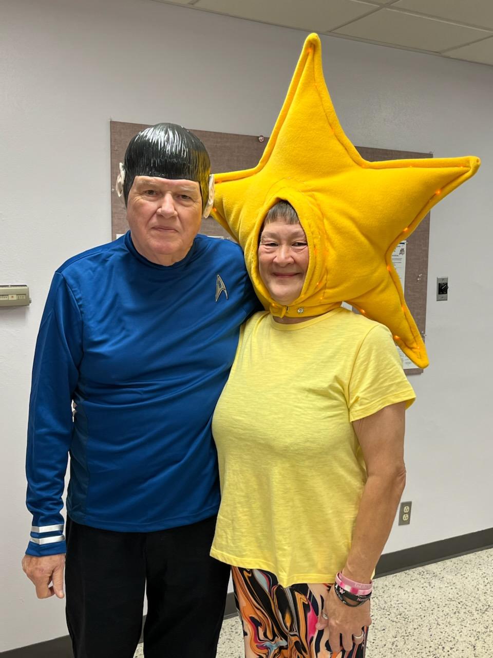 Two people pose indoors, one in a blue shirt and one in a yellow star costume, both smiling.