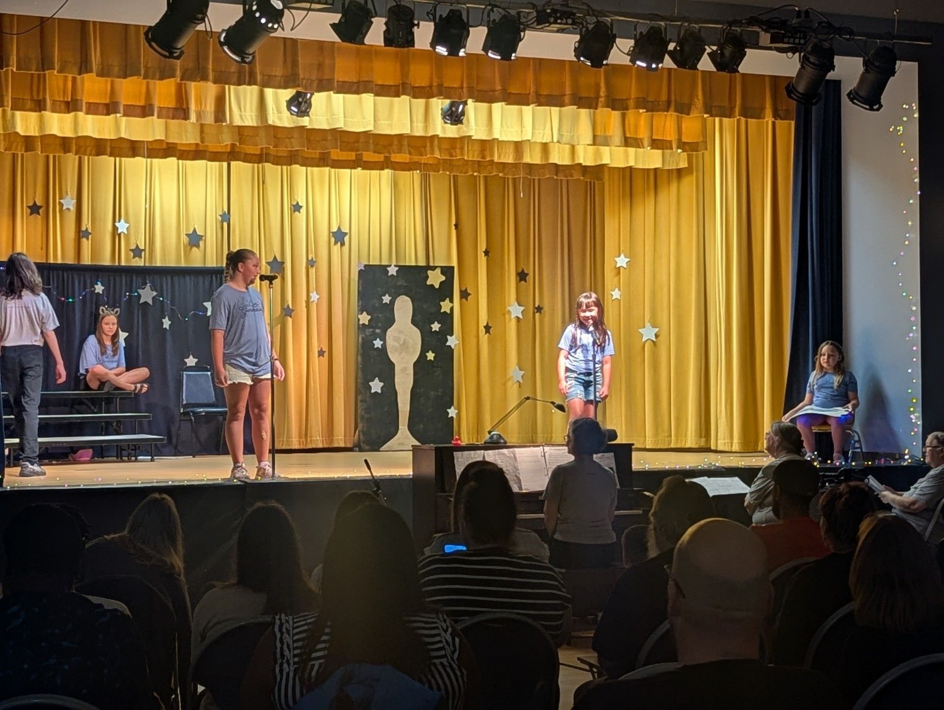 Stage performance with two people under gold curtains, audience seated in front, and musicians at the sides.