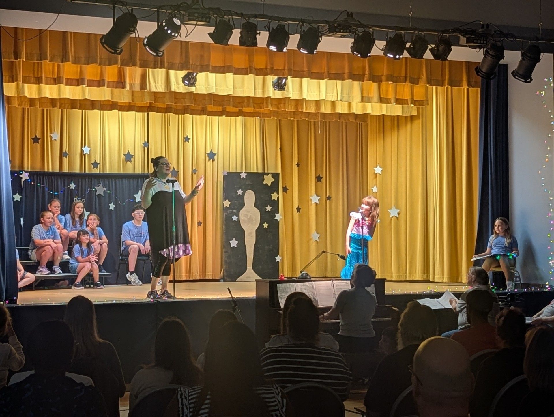 Stage performance with children in costumes on a gold-curtained theater stage, audience seated in front