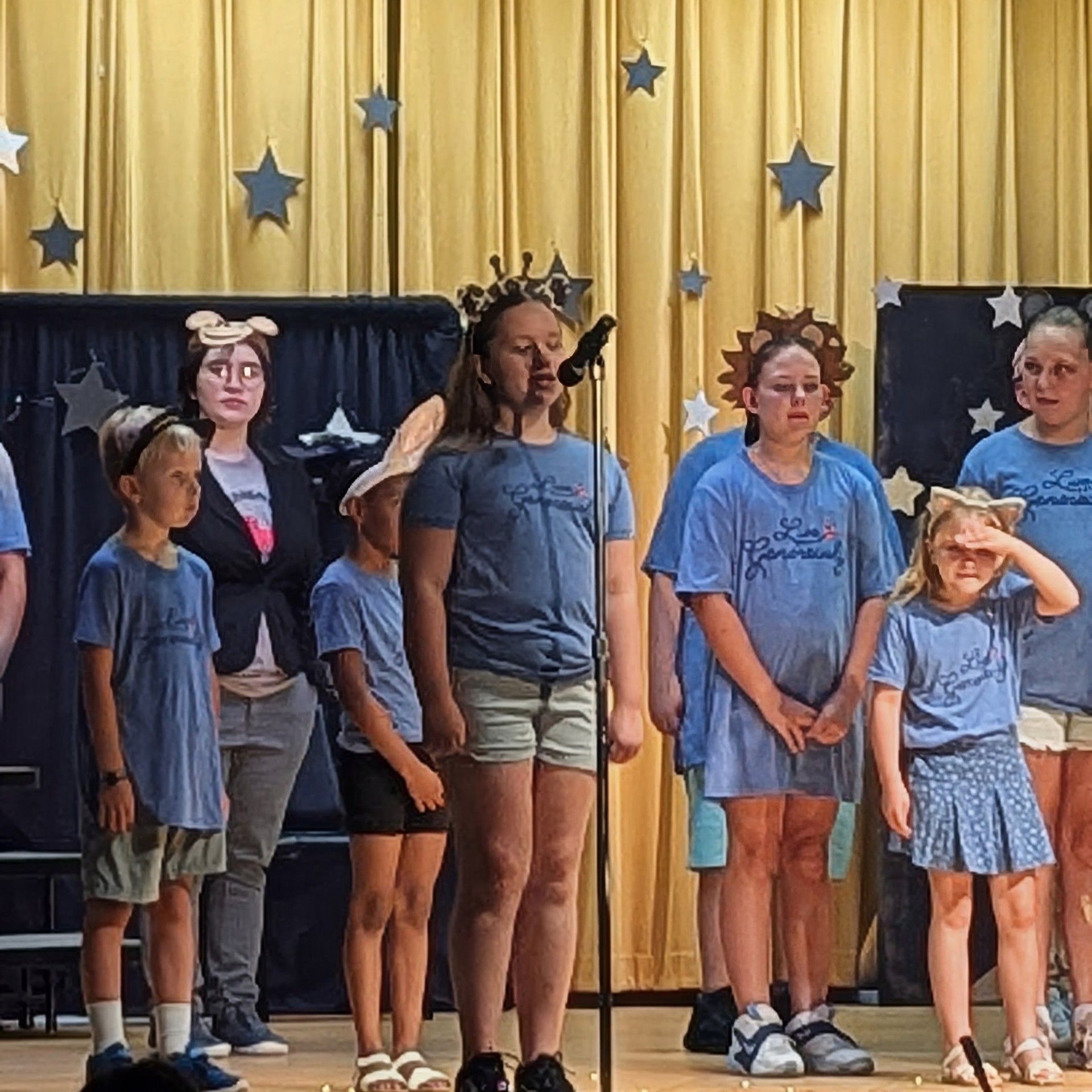 Children on a stage in blue shirts and shorts, standing before starry curtains and a gold backdrop.
