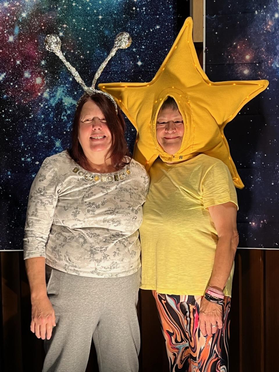 Two people posing indoors in costumes, one in a gold star hat and yellow shirt, against a starry backdrop.