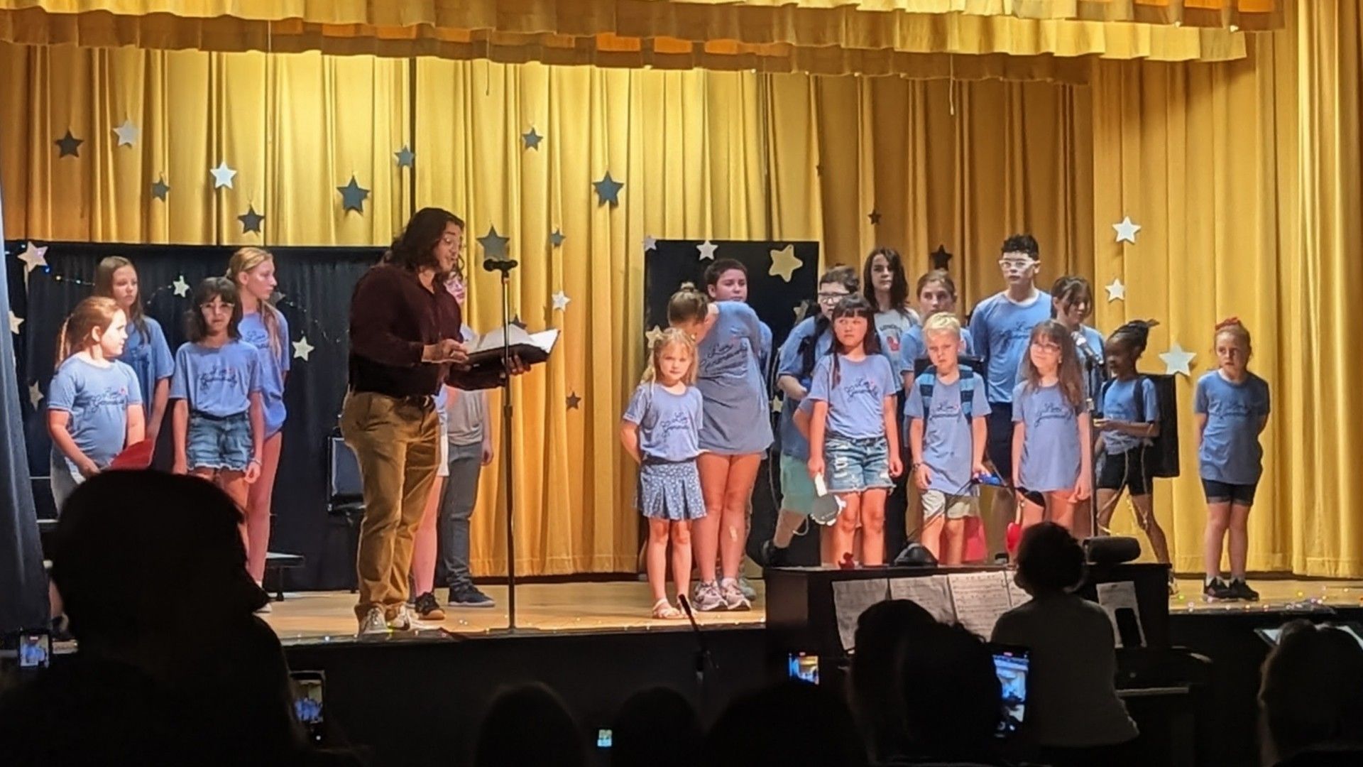 Children in blue shirts sing on a stage with a conductor in front of gold curtains