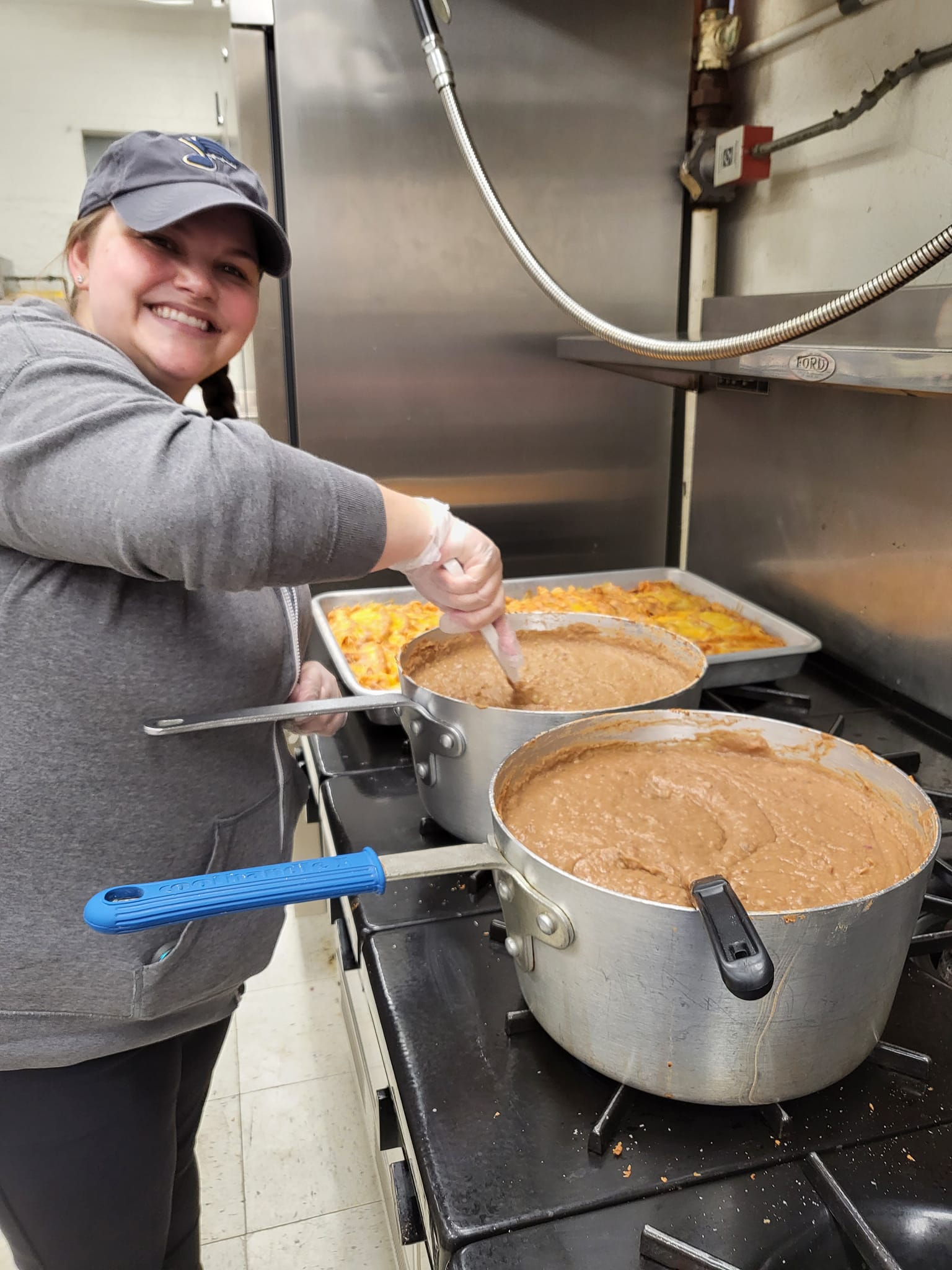 Person in a kitchen stirring a large pot of stew beside trays of food.