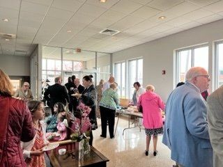 People mingling in a bright lobby, some holding flowers, near large windows and seated tables.