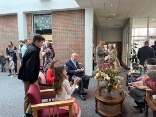 People seated and standing in a bright lobby around a wicker table with flowers and snacks