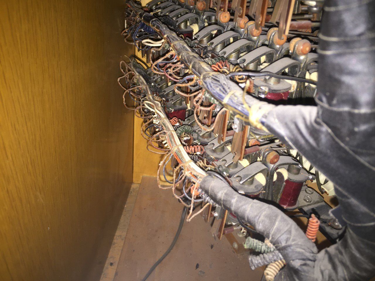 Hands inspecting a dense bundle of wires and components inside a wooden cabinet.