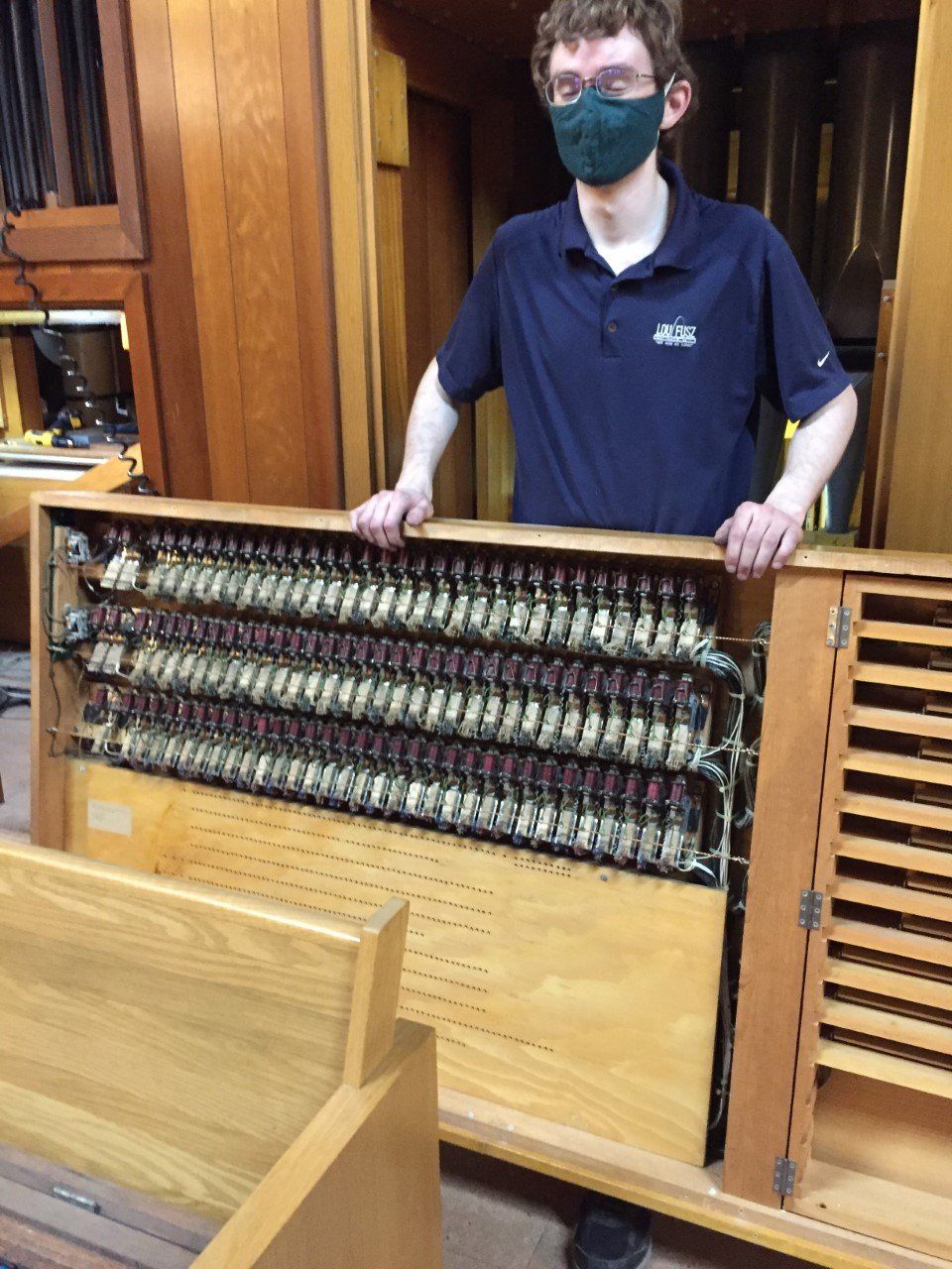 Man standing indoors beside a large wooden mechanical keyboard mechanism with rows of metal parts
